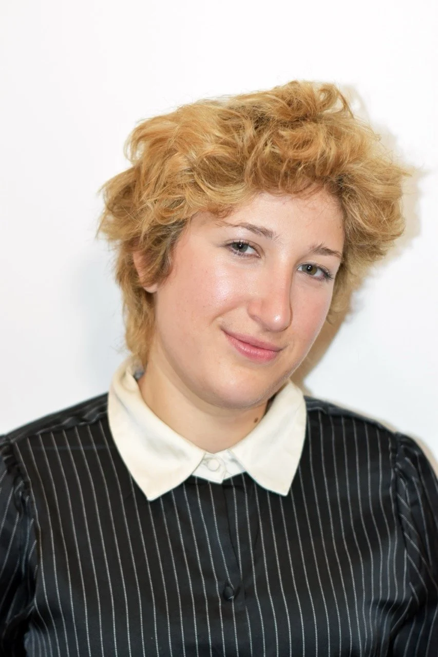Young woman with short, curly, blonde hair smiling, wearing a black pinstripe shirt with a white collar, against a white background.