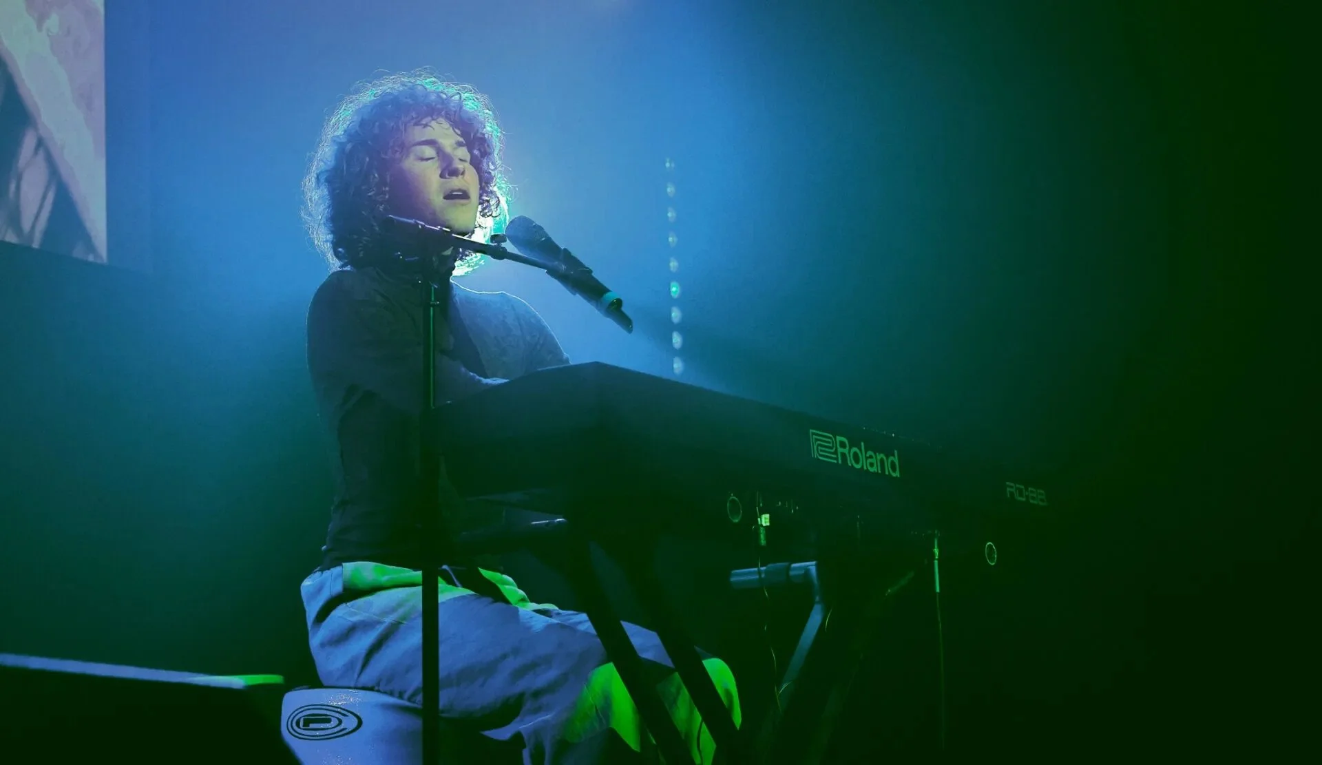 A woman with curly hair is performing on stage, playing a Roland electronic keyboard and singing into a microphone, with blue and green stage lighting.