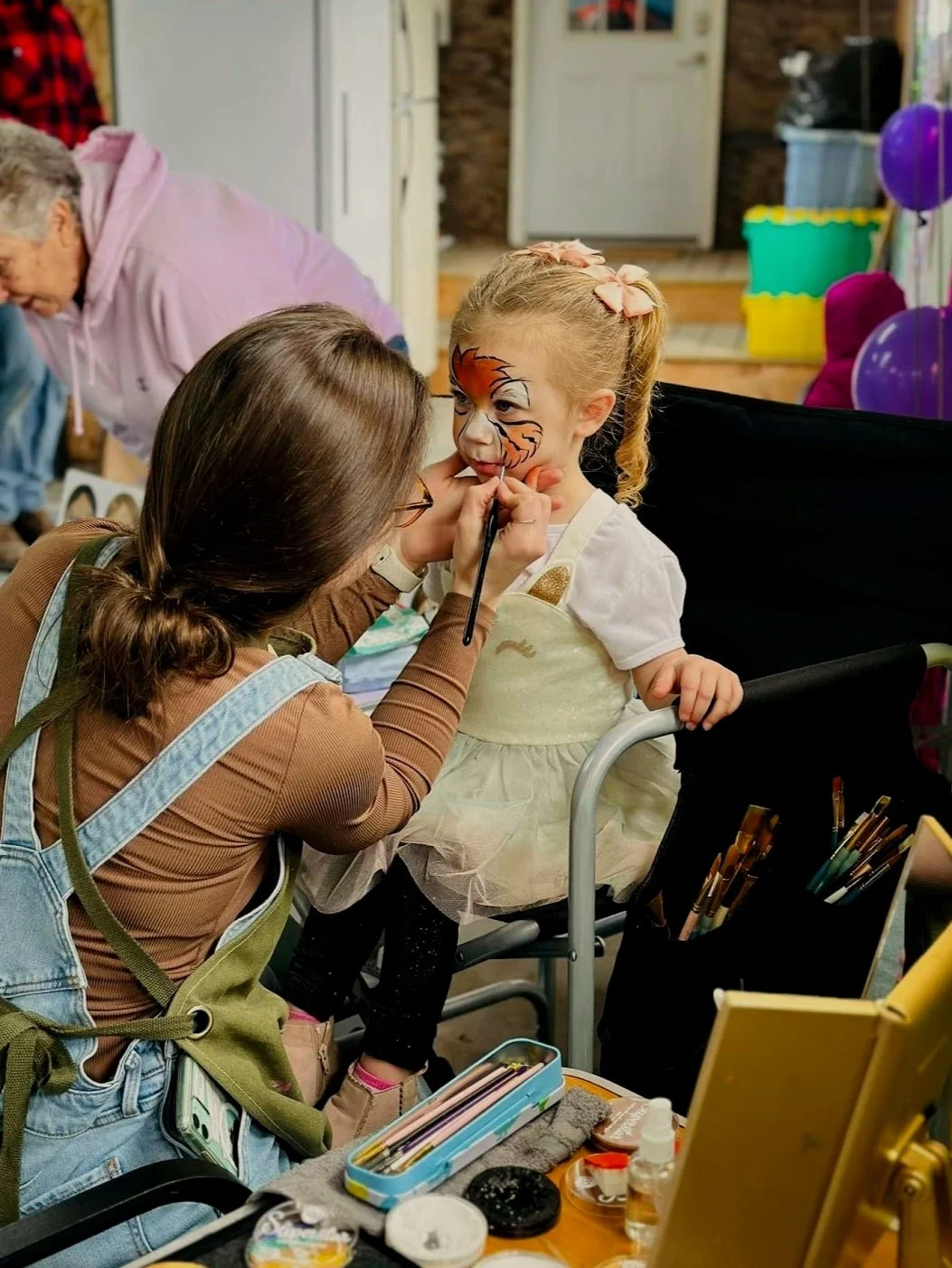 A young girl with a tiger face painted on her face sits in a chair while a face painter carefuly applies details at a party or event, with colorful balloons and decorations in the background.