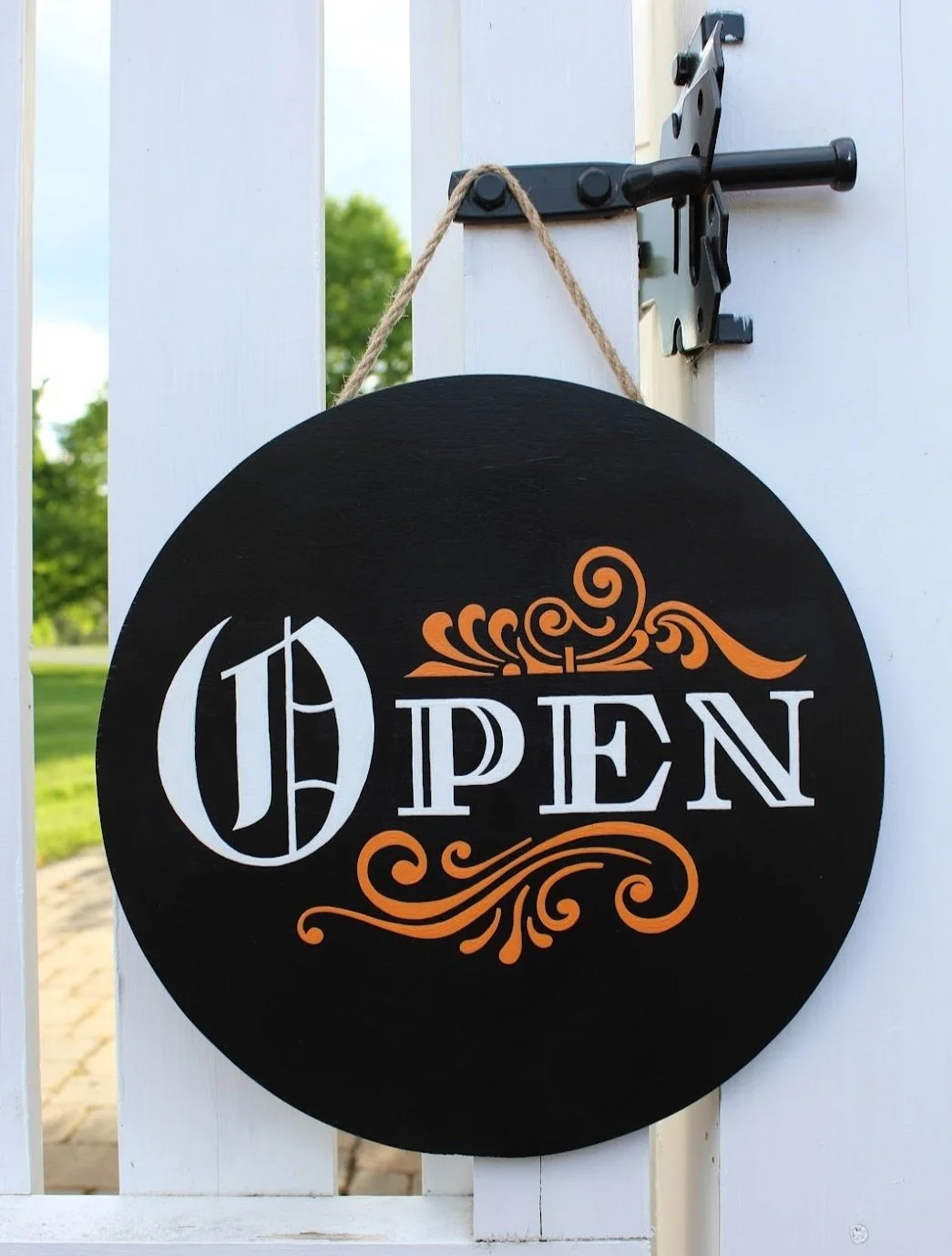Black circular sign hanging on a white gate that reads 'OPEN' in stylized white and orange lettering.