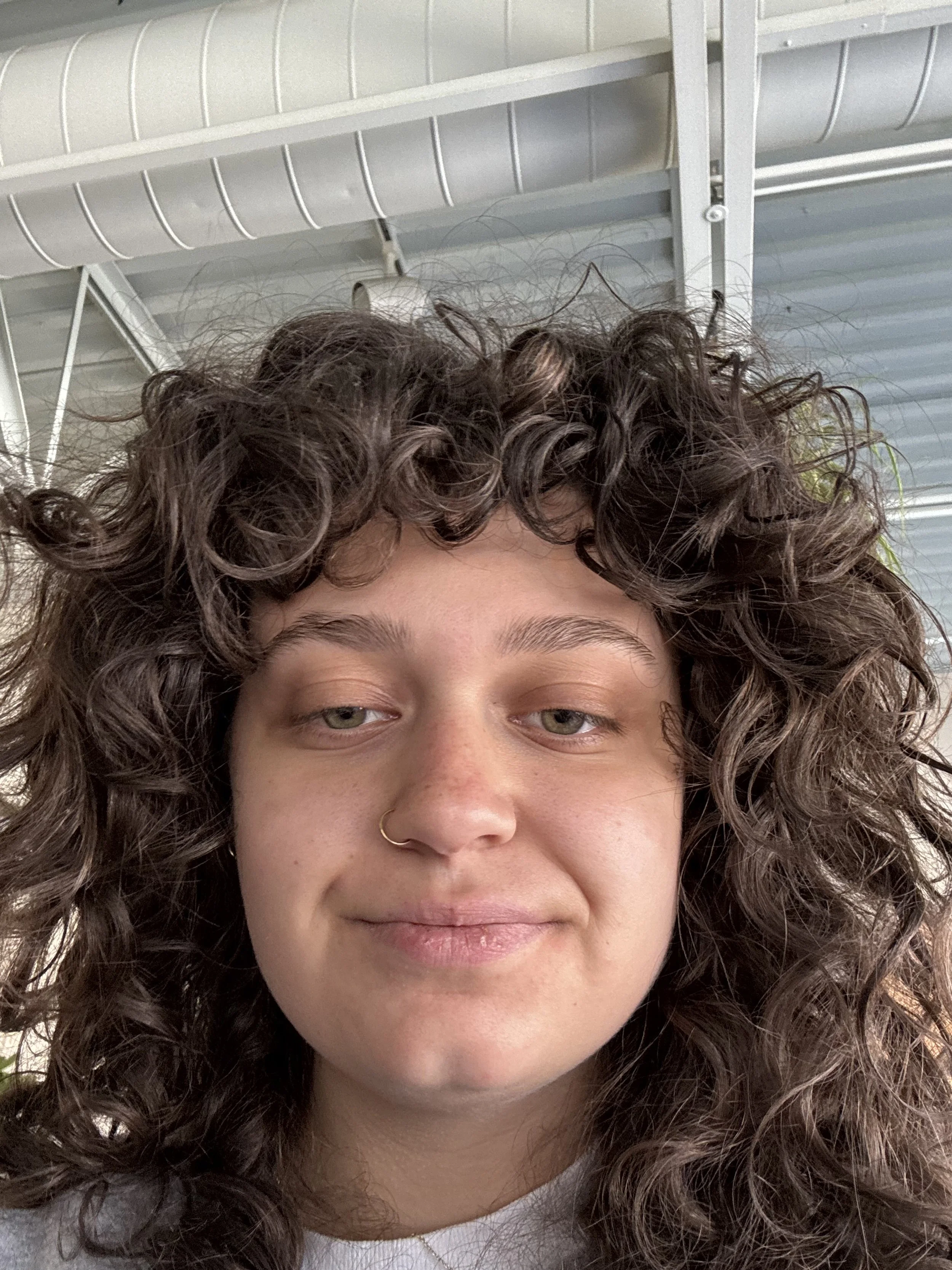 A young woman with curly brown hair, light skin, and a nose piercing, smiling softly, indoors with visible metal beams and ceiling vents in the background.