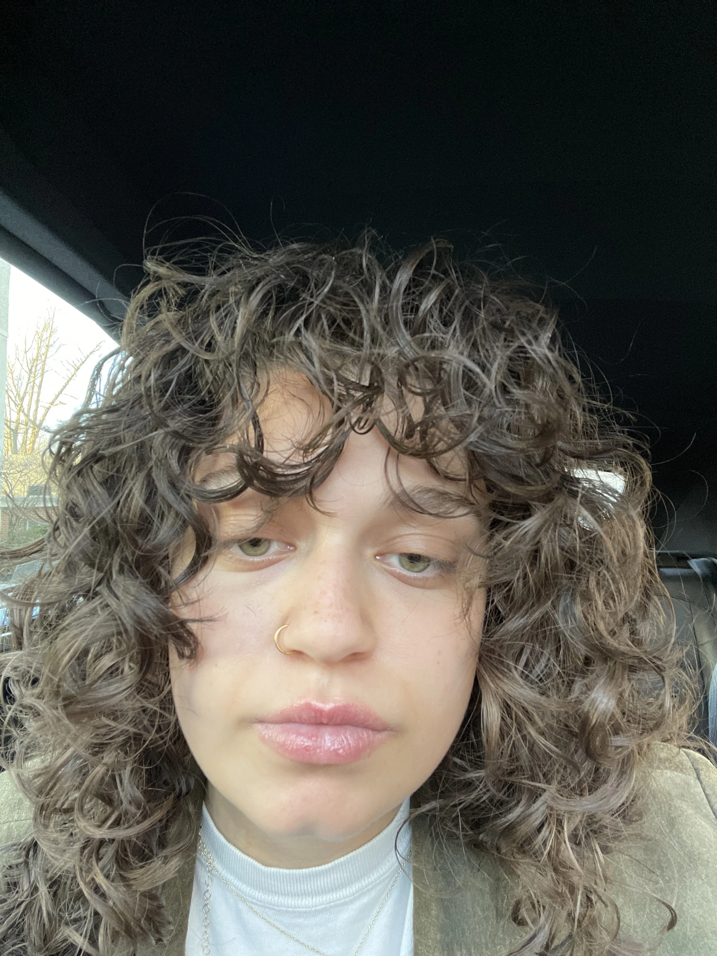 Close-up of a woman with curly hair, green eyes, wearing a gold nose ring, a white turtleneck, and a gray blazer inside a vehicle.