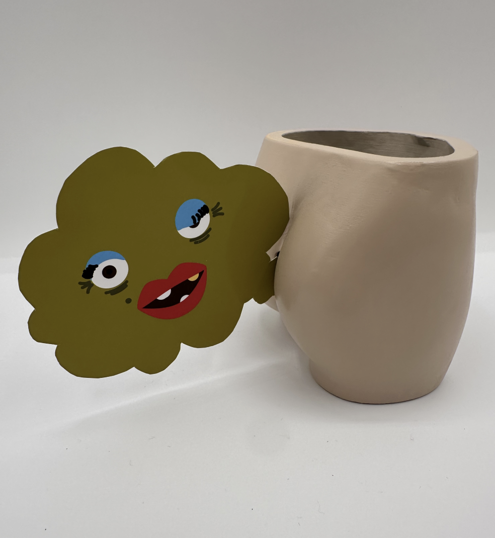 A beige, irregularly shaped ceramic vase beside a cartoonish paper cutout of a smiling, winking face with blue eyes, red lips, and black eyelashes on a light grey background.