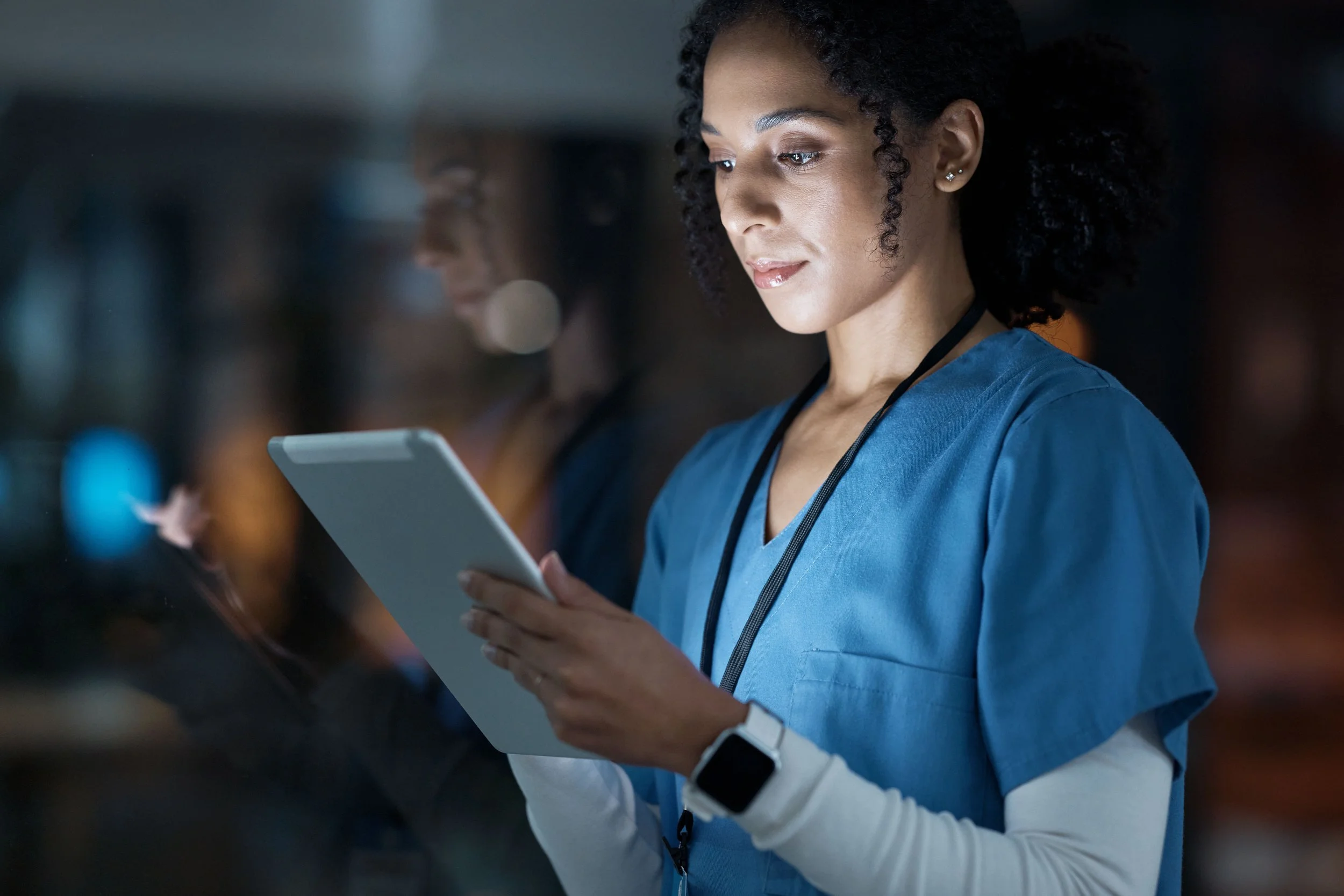 Female healthcare worker in blue scrubs and a smartwatch looking at a tablet in a clinical setting.