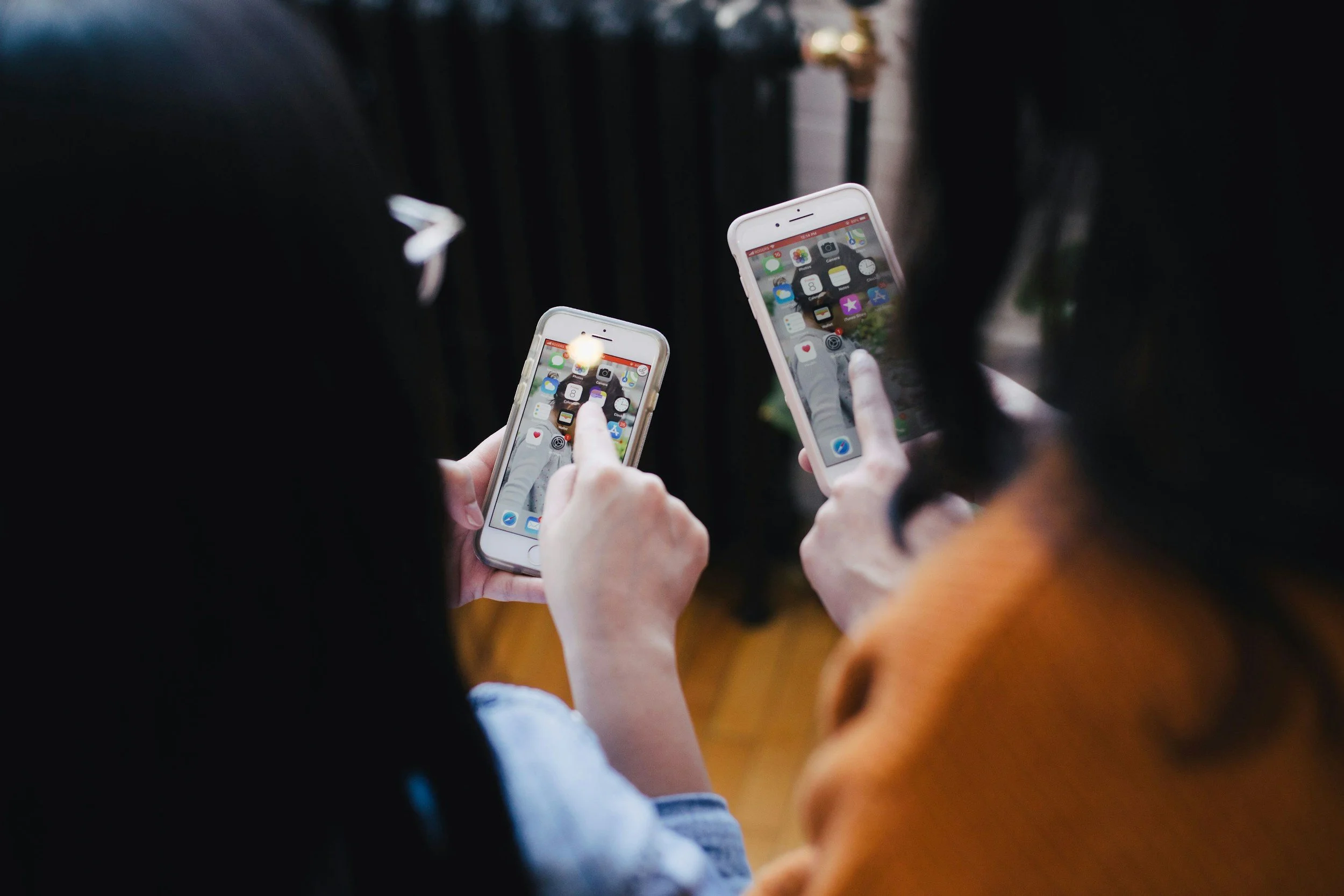 Two women are looking at their smartphones, both screens showing the same app icons. One woman has dark hair and glasses, and the other has dark hair and an orange shirt.