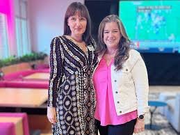 Two women standing together indoors, smiling at the camera. The woman on the left is wearing a patterned dress, and the woman on the right is wearing a white jacket over a pink top. Behind them is a pink and teal seating area and a screen displaying a presentation.