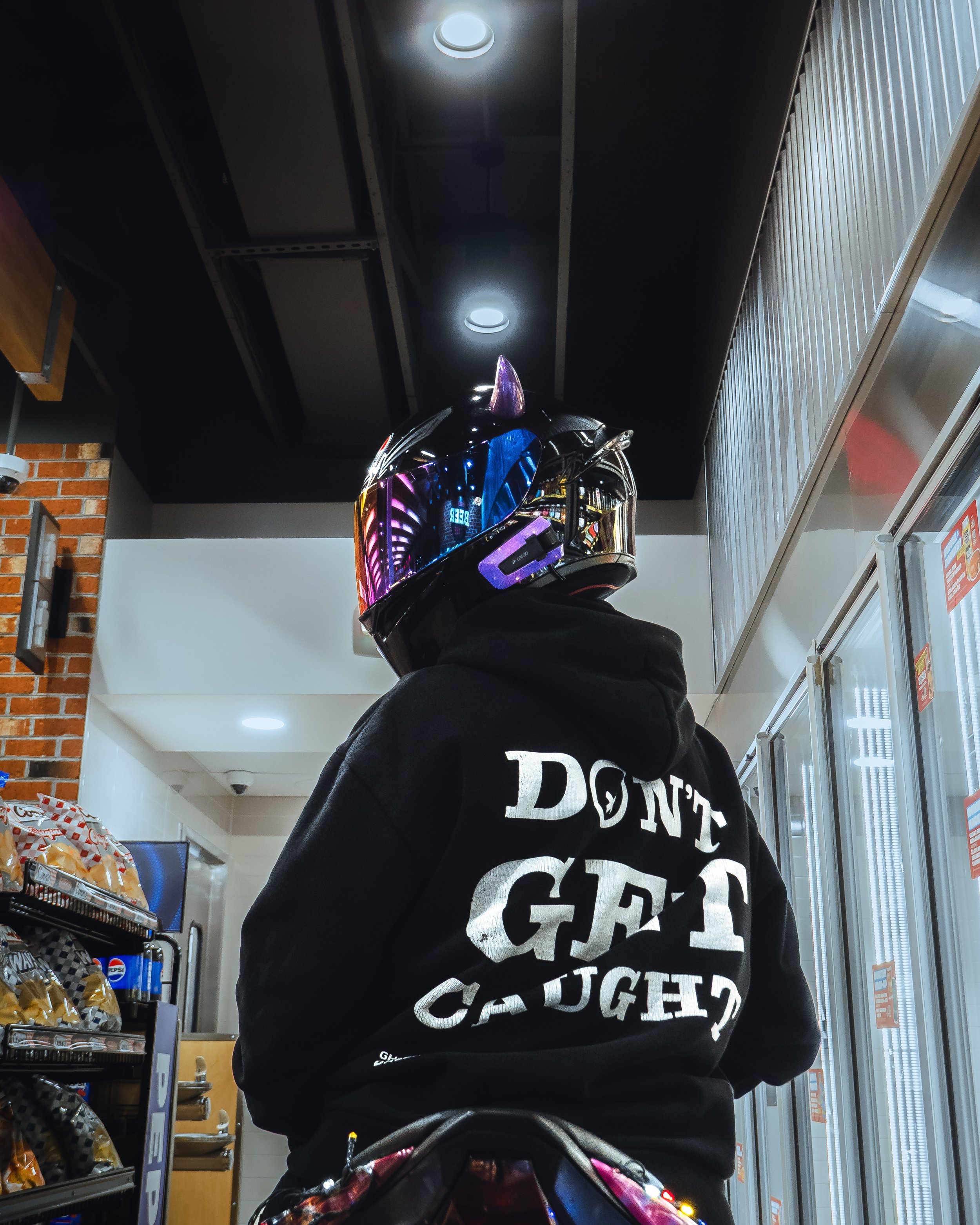 Person wearing a shiny, reflective helmet with purple unicorn horn detail, a black hoodie with white text, and sitting on a scooter inside a convenience store.