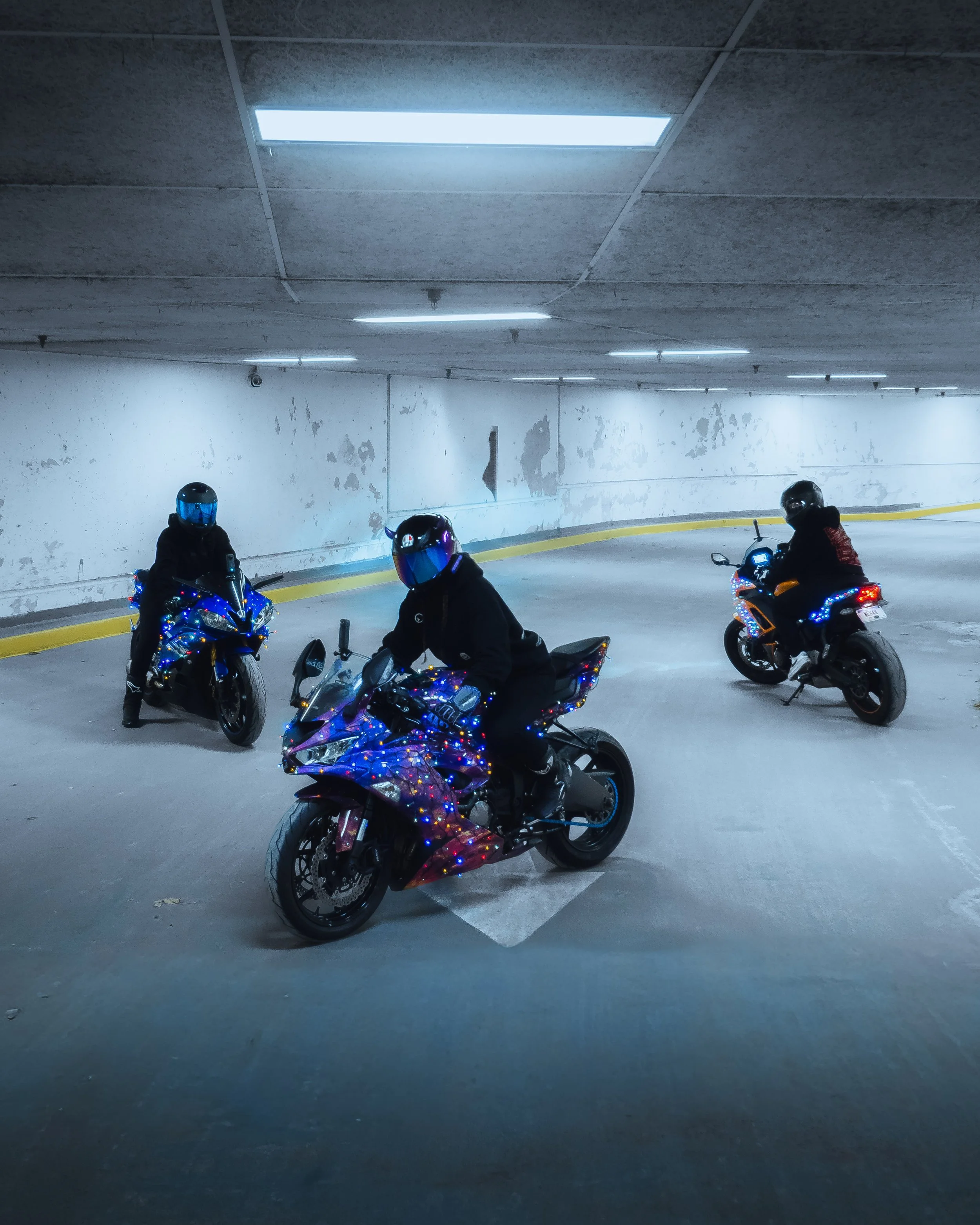 Three motorcyclists wearing helmets and black jackets in an underground parking garage, decorated with colorful string lights.