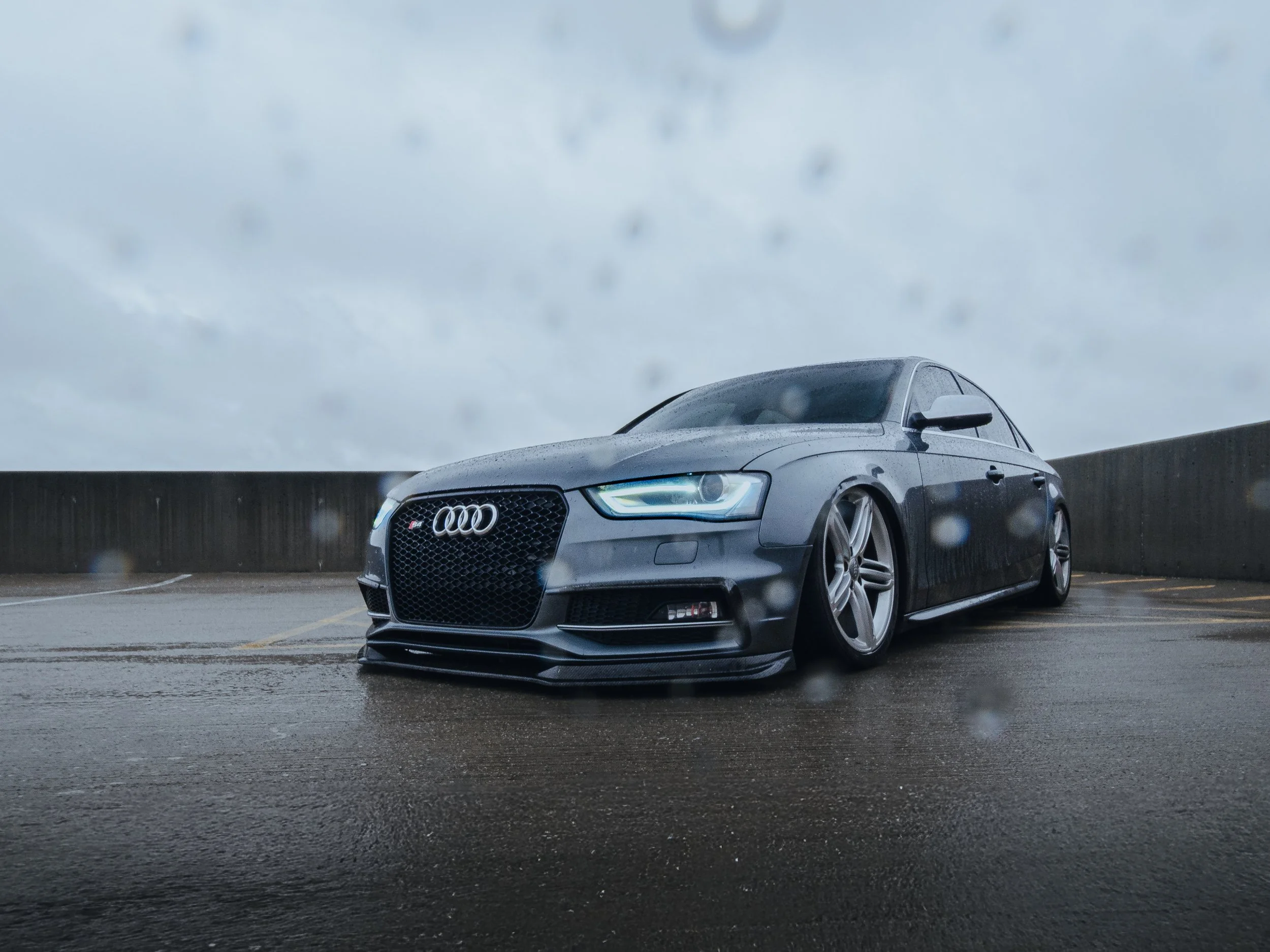 Gray Audi sedan parked on a wet parking lot during rainy weather with raindrops on the camera lens and overcast sky.