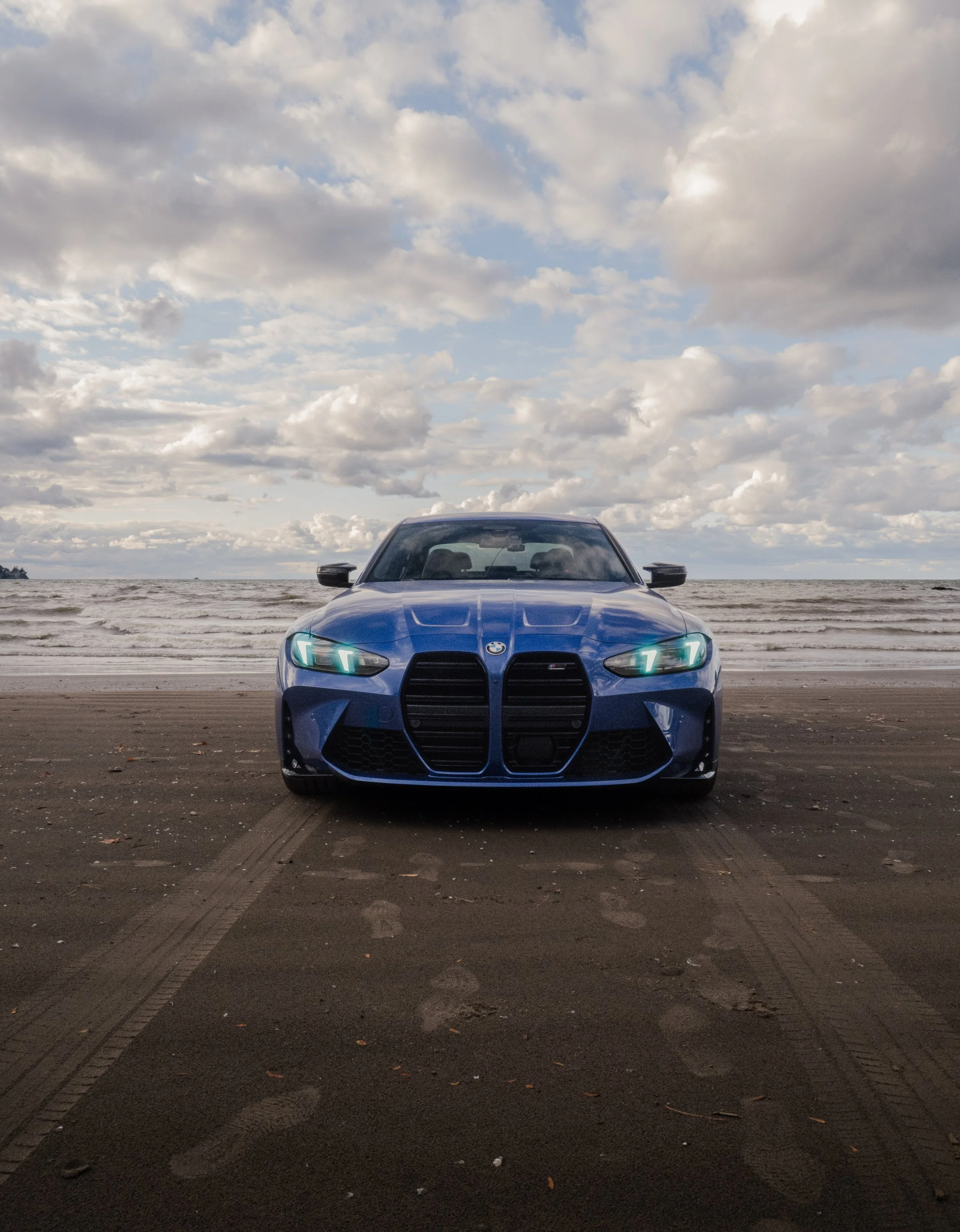 A blue BMW car parked on a sandy beach facing the ocean, with a cloudy sky overhead.