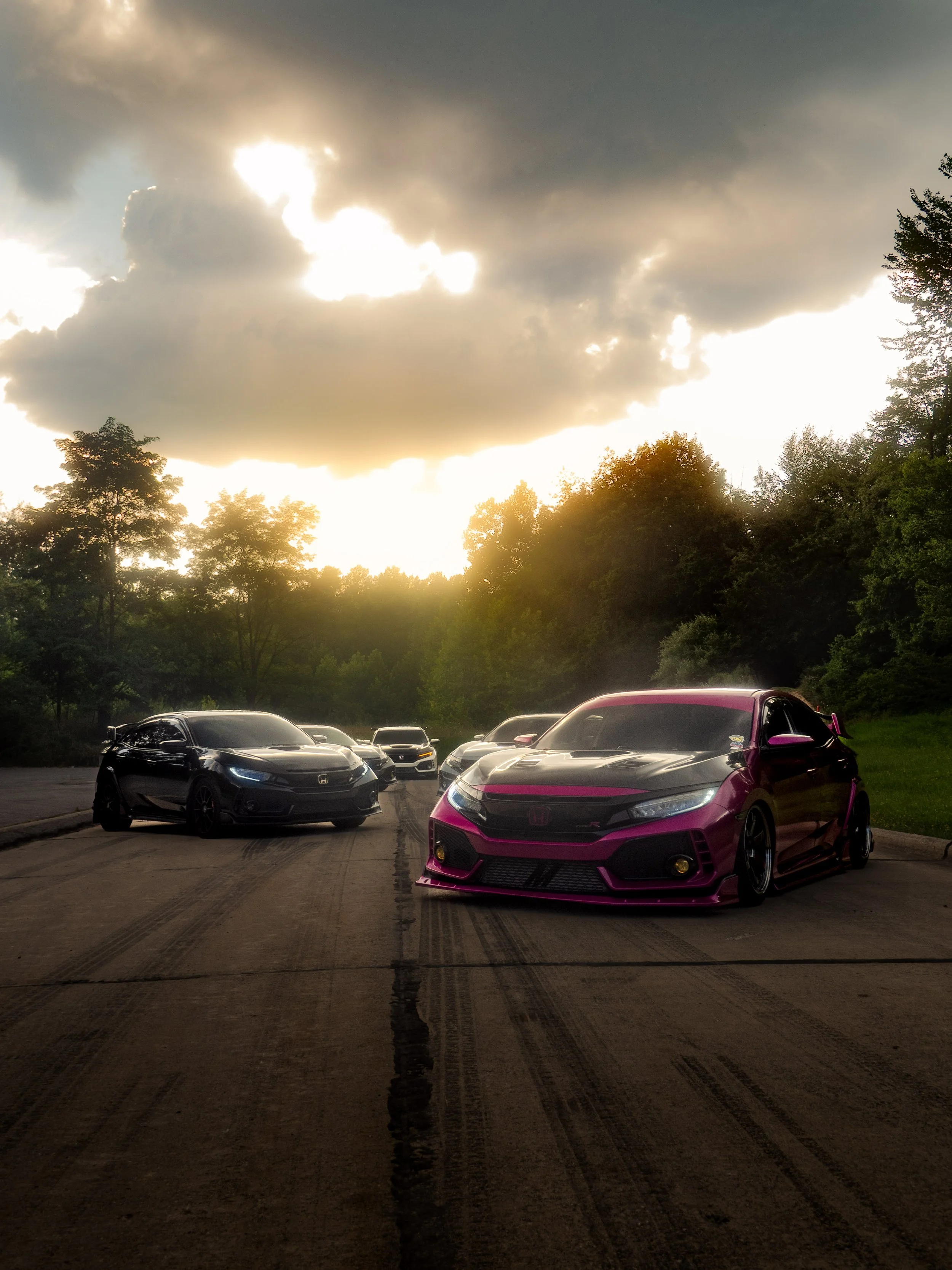 Several cars parked on a concrete surface with tire marks, trees and a cloudy sunset sky in the background.