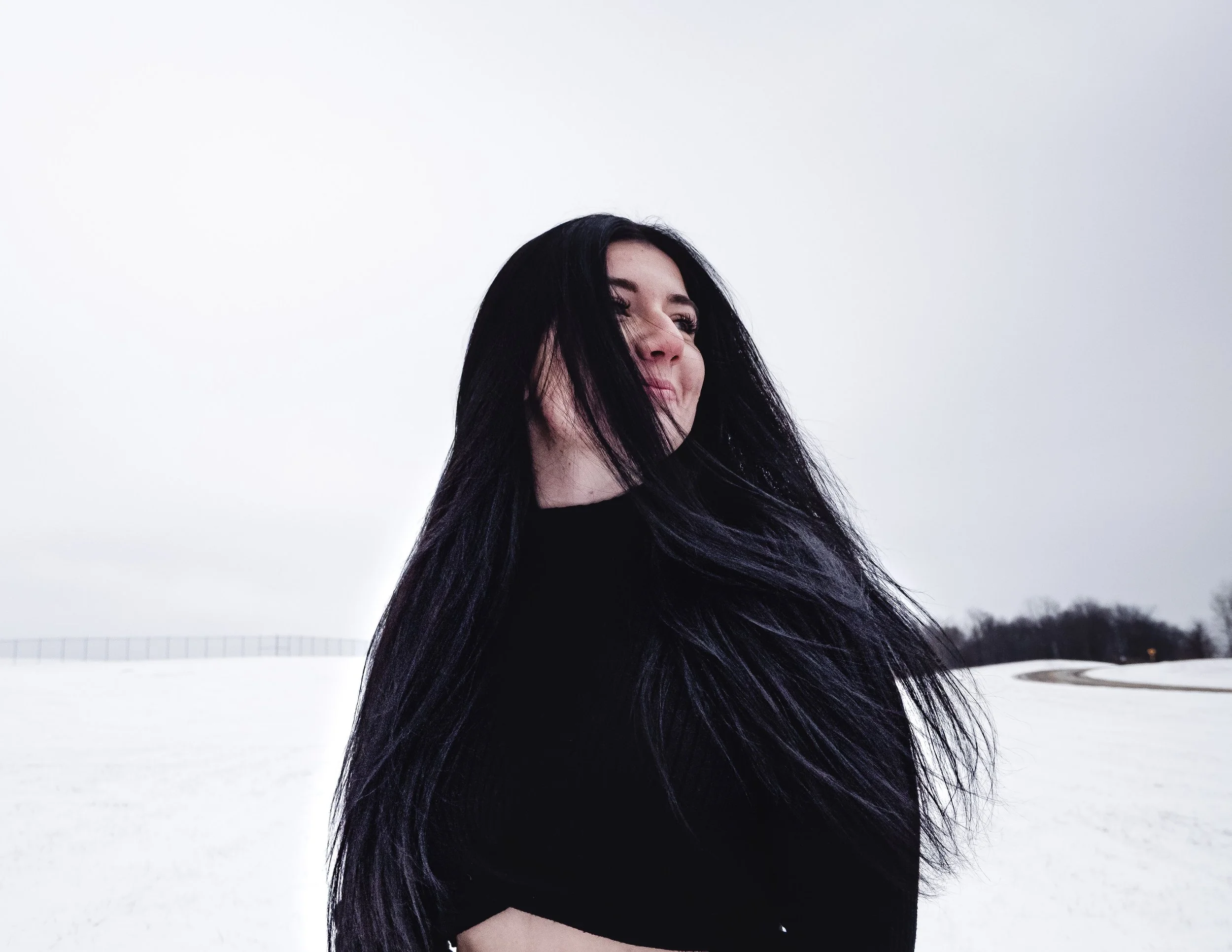 A woman with long black hair wearing a black top standing in a snowy landscape outdoors with an overcast sky.