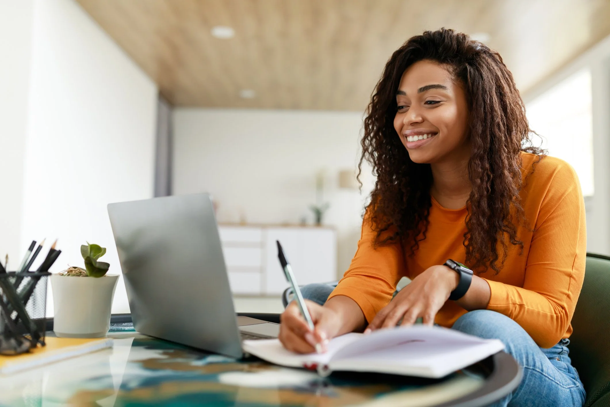 Young woman with curly hair and an orange shirt smiling while working at a desk.