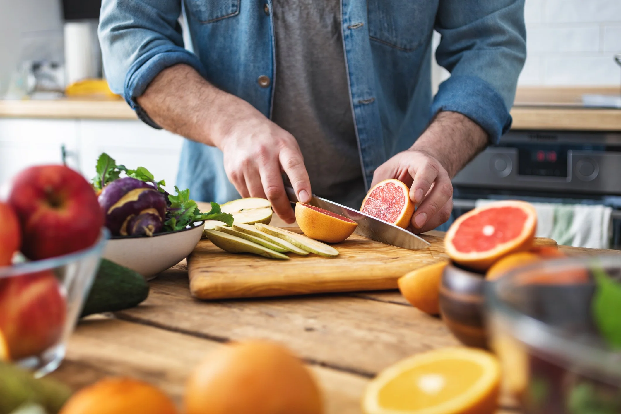 Person slicing a grapefruit in a modern kitchen. Various fresh fruits on a wooden table, including apples, pears, and grapes.