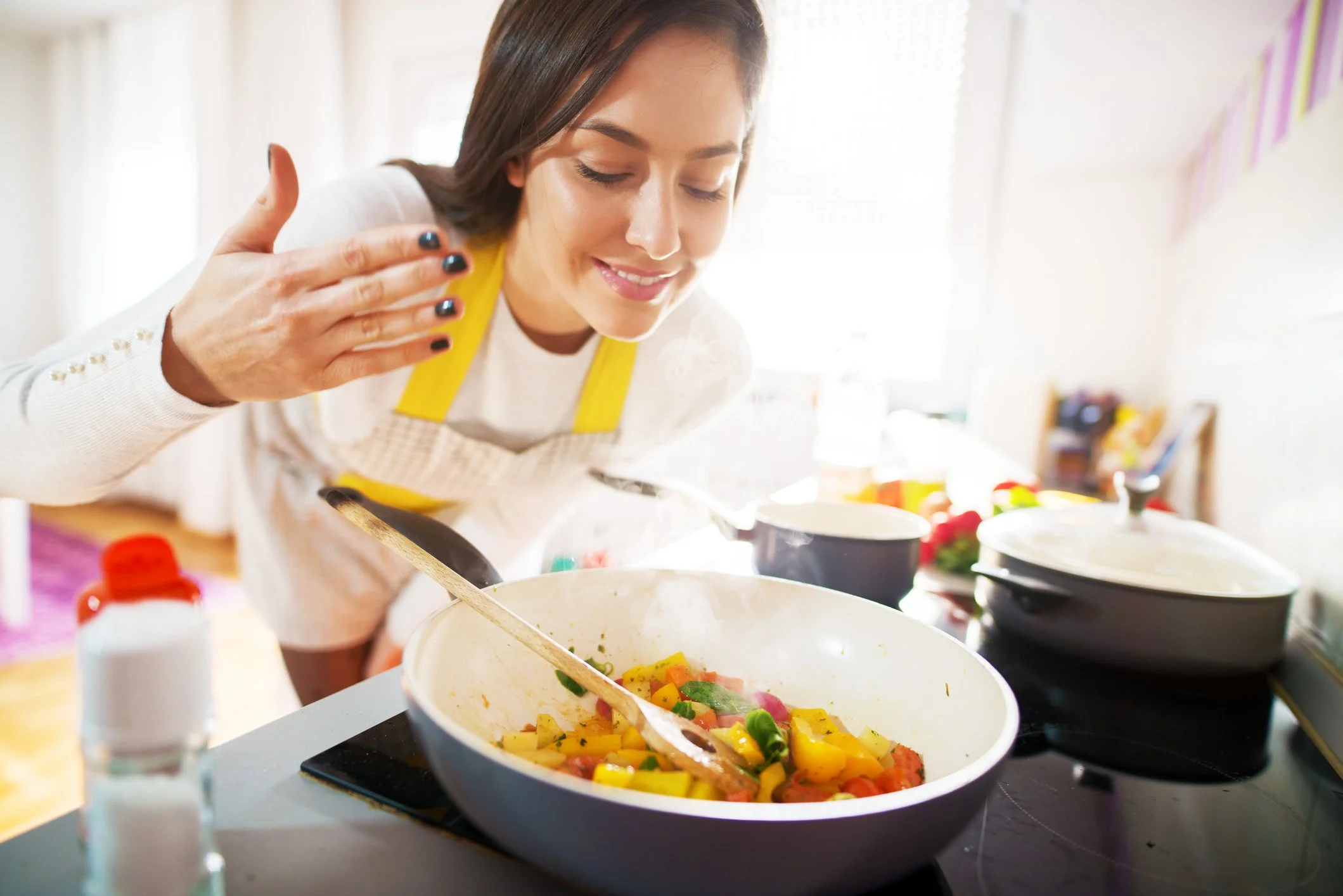 Woman with dark hair and yellow apron preparing a colorful salad or dish in a kitchen with natural light; various bowls and cooking ingredients on the table.