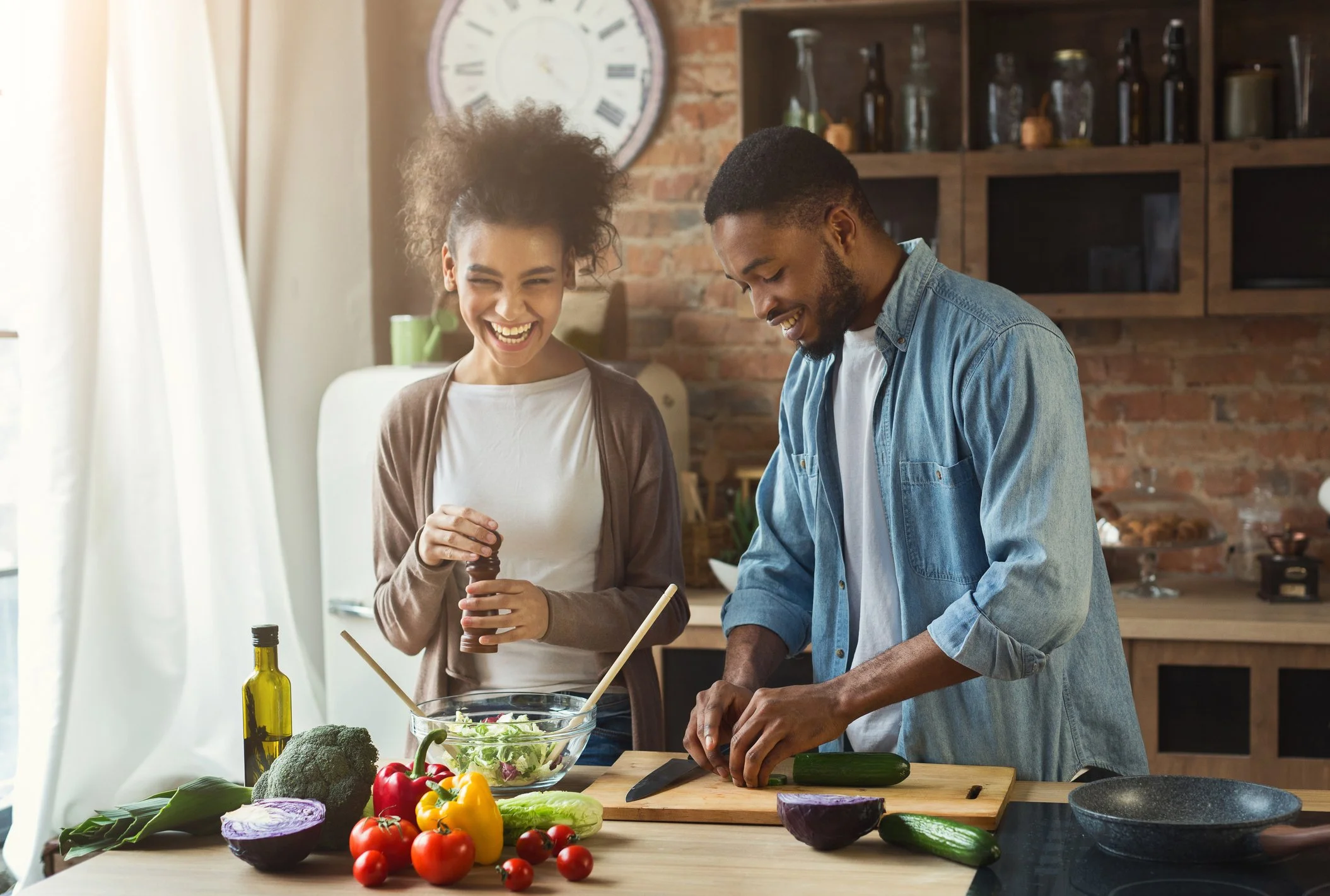 A woman and a man cooking together in a kitchen with fresh vegetables, a glass bowl of salad, and utensils on the wooden table.