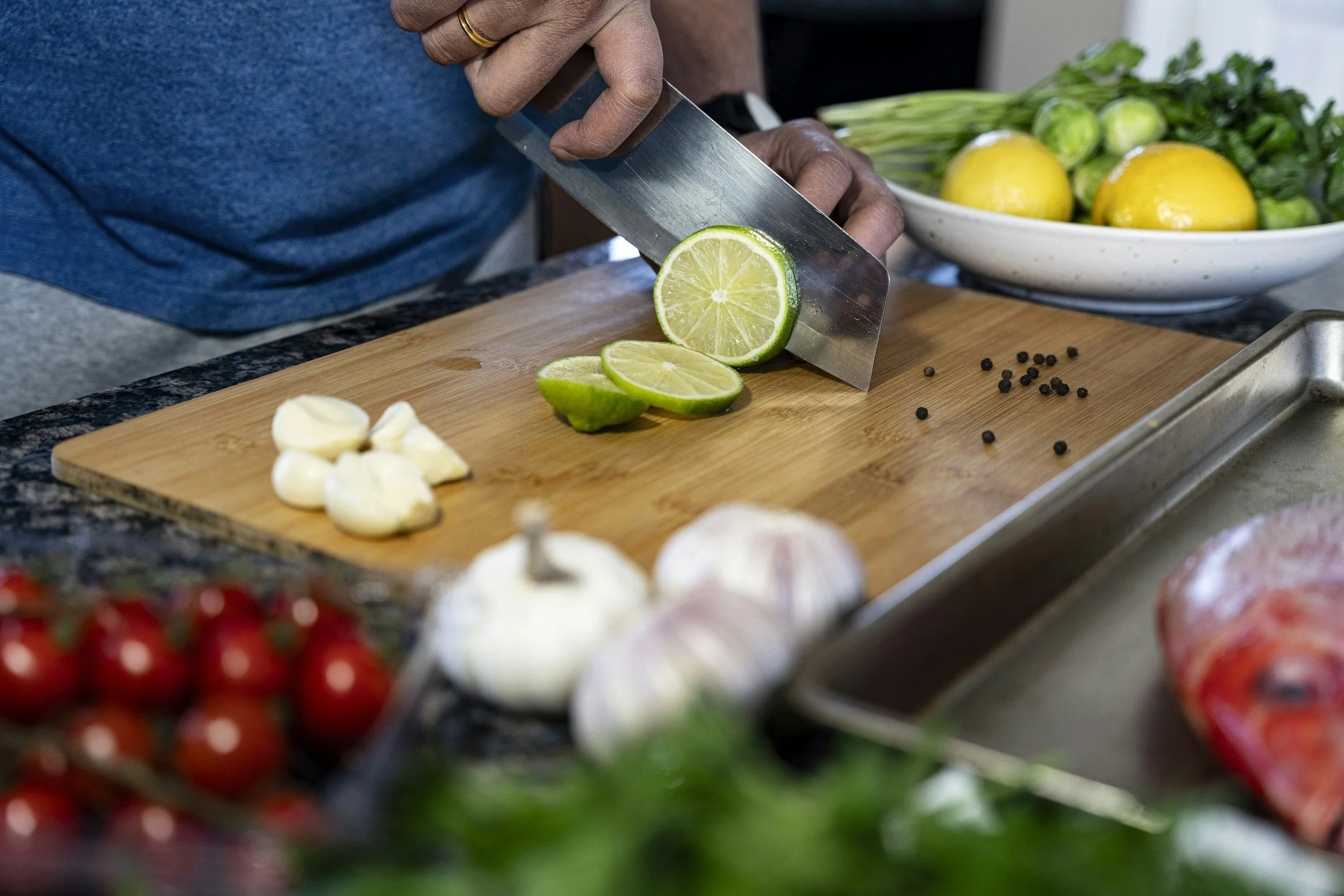 A person slicing a lime on a wooden cutting board, with garlic cloves nearby and a bowl of lemons and greens in the background.