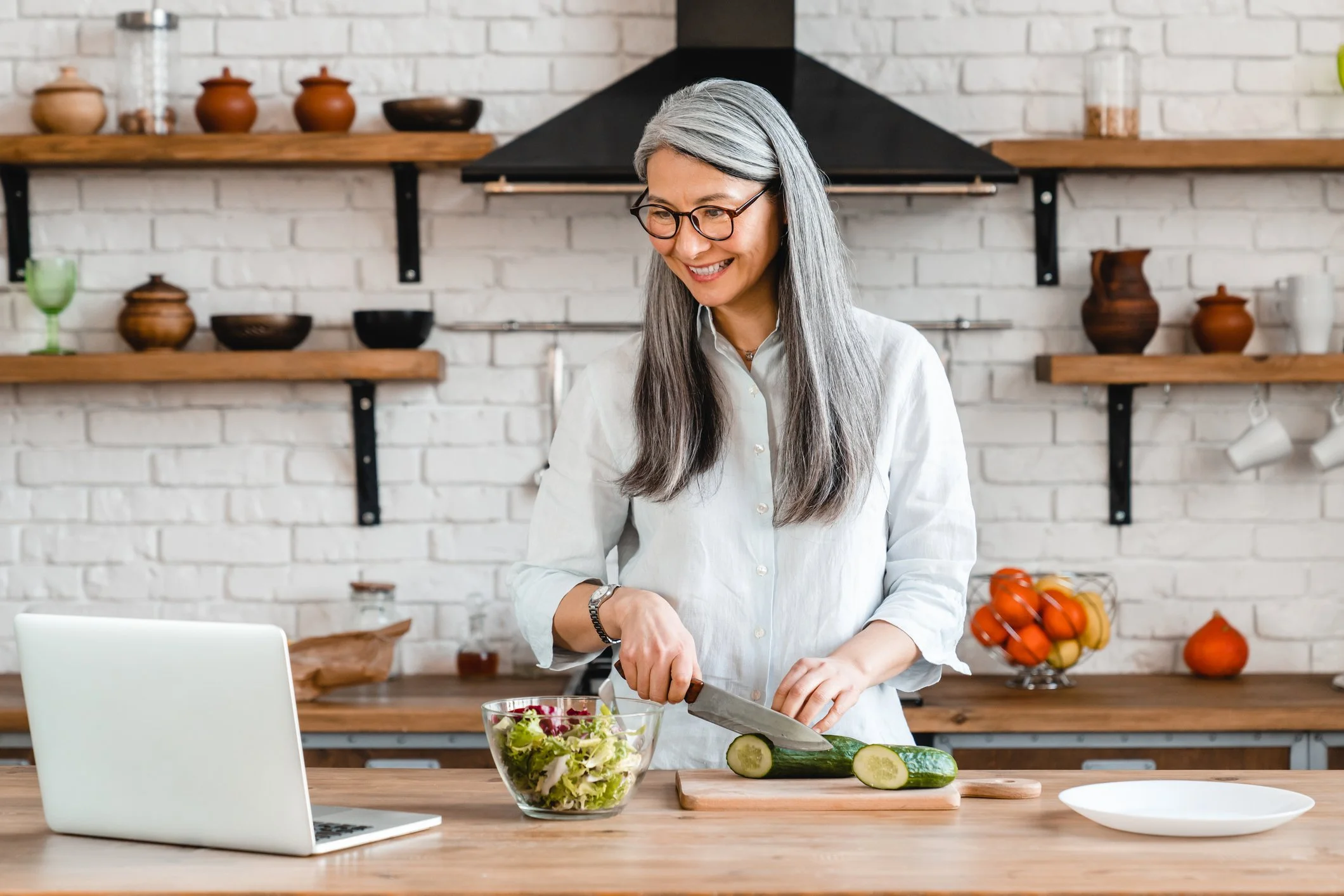 A woman with long gray hair and glasses chopping cucumbers and preparing a salad in a modern kitchen with open wooden shelves and white brick wall.