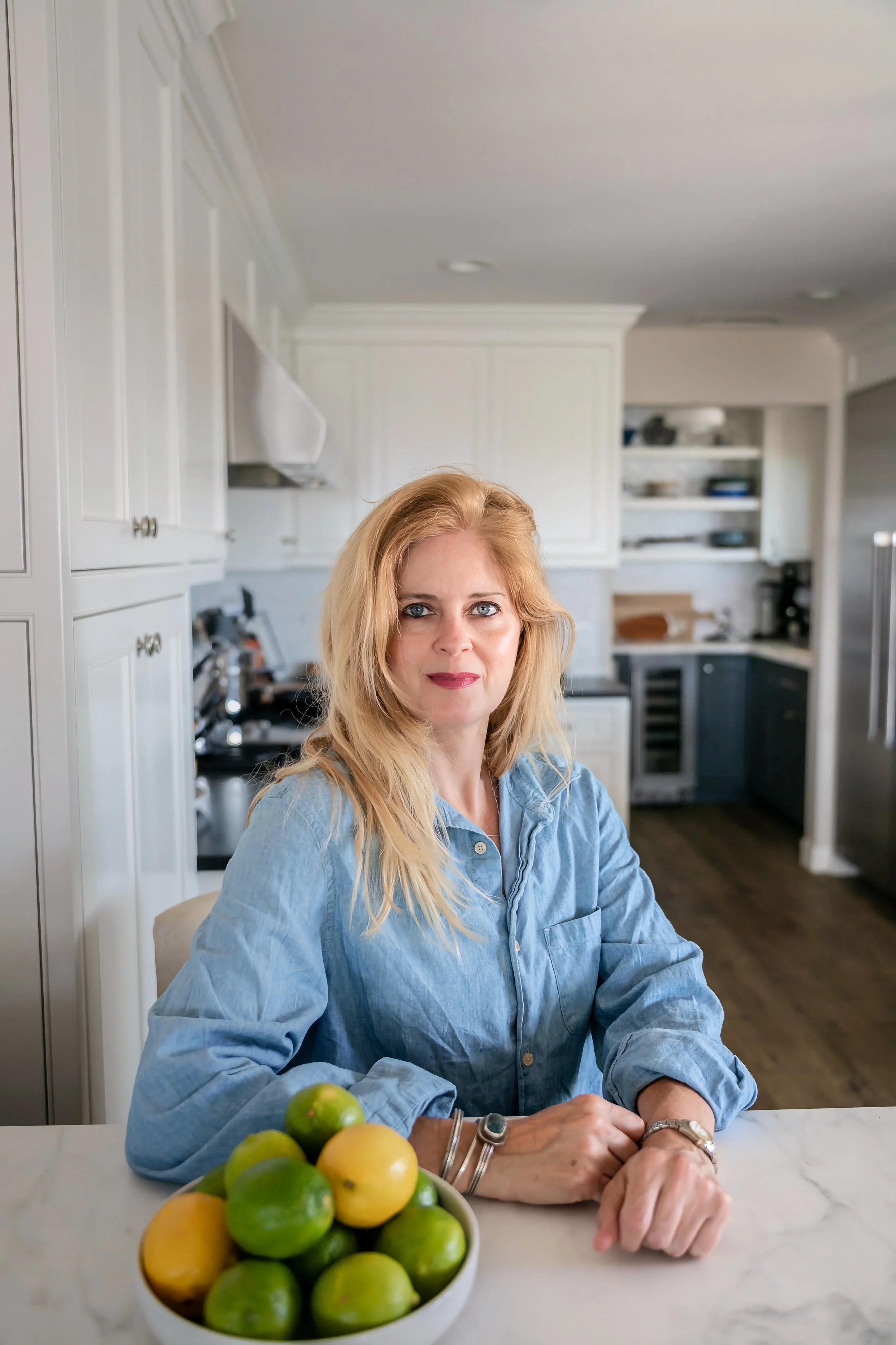 A woman with blonde hair and blue eyes, wearing a light blue denim shirt, sits at a kitchen counter with a bowl of lemons and limes in front of her, in a bright white kitchen with open shelving and cabinetry.