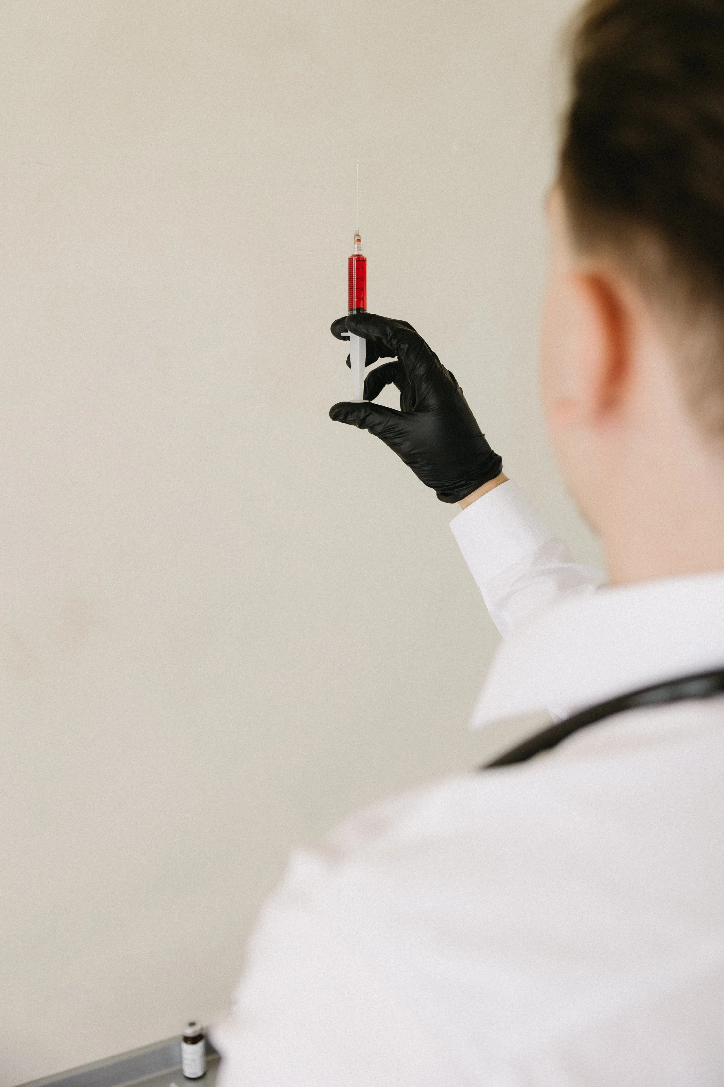 A medical professional wearing black gloves and a white coat holding a syringe with red liquid.