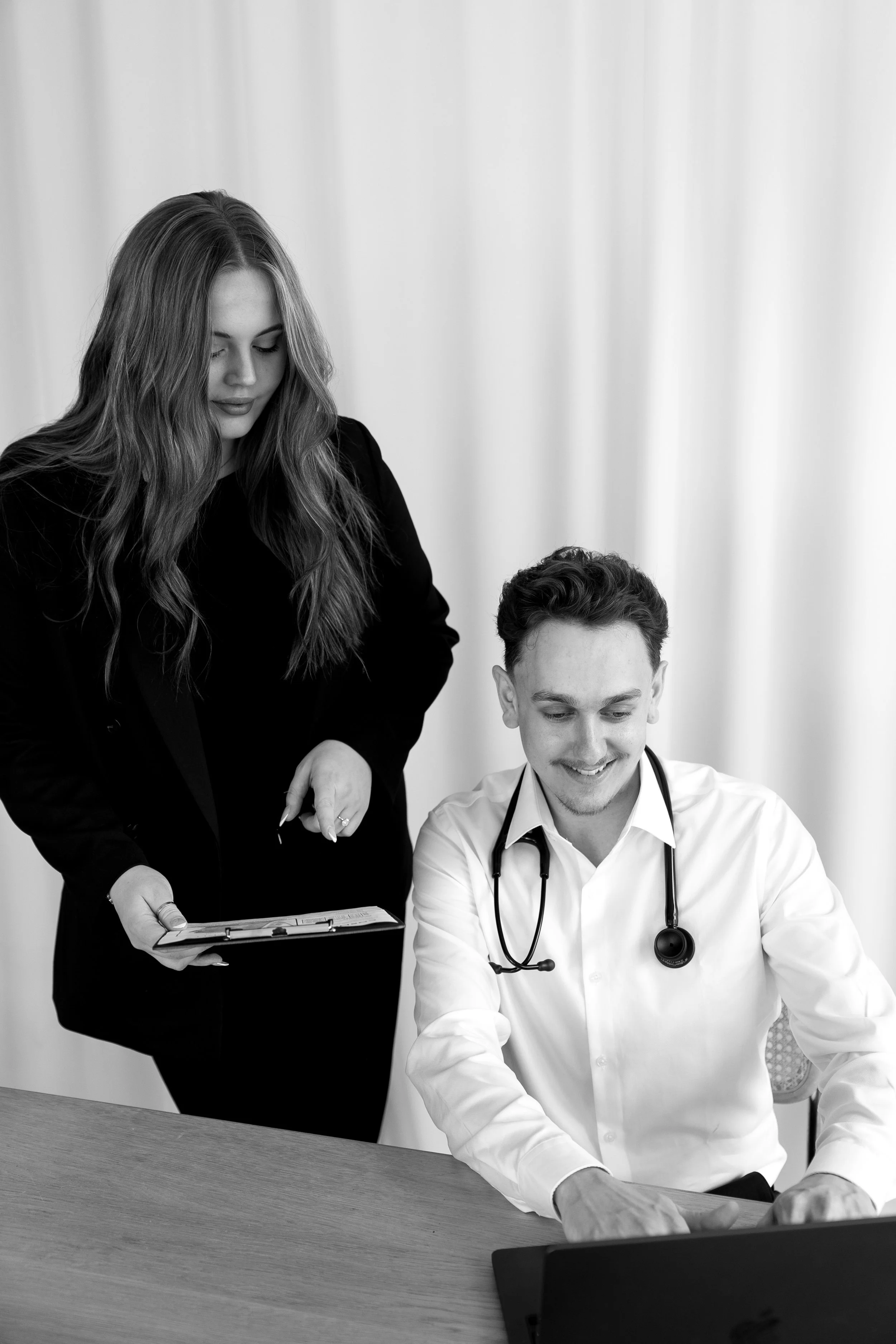 A woman and a man, who is a doctor with a stethoscope, looking at a laptop and a clipboard in a professional setting.