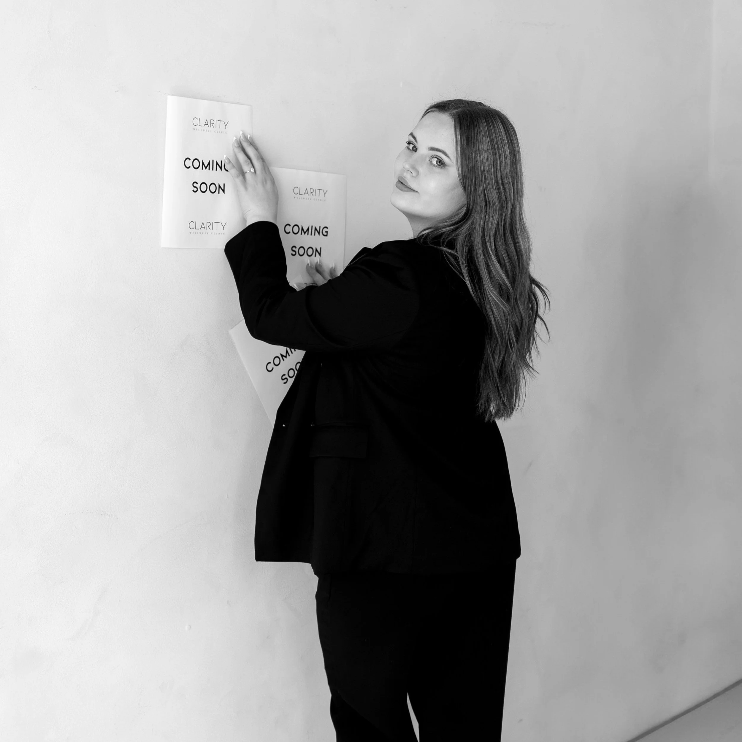 A woman with long hair in a black suit sticking posters on a white wall, the posters read 'COMING SOON' with the word 'CLARITY' at the top.
