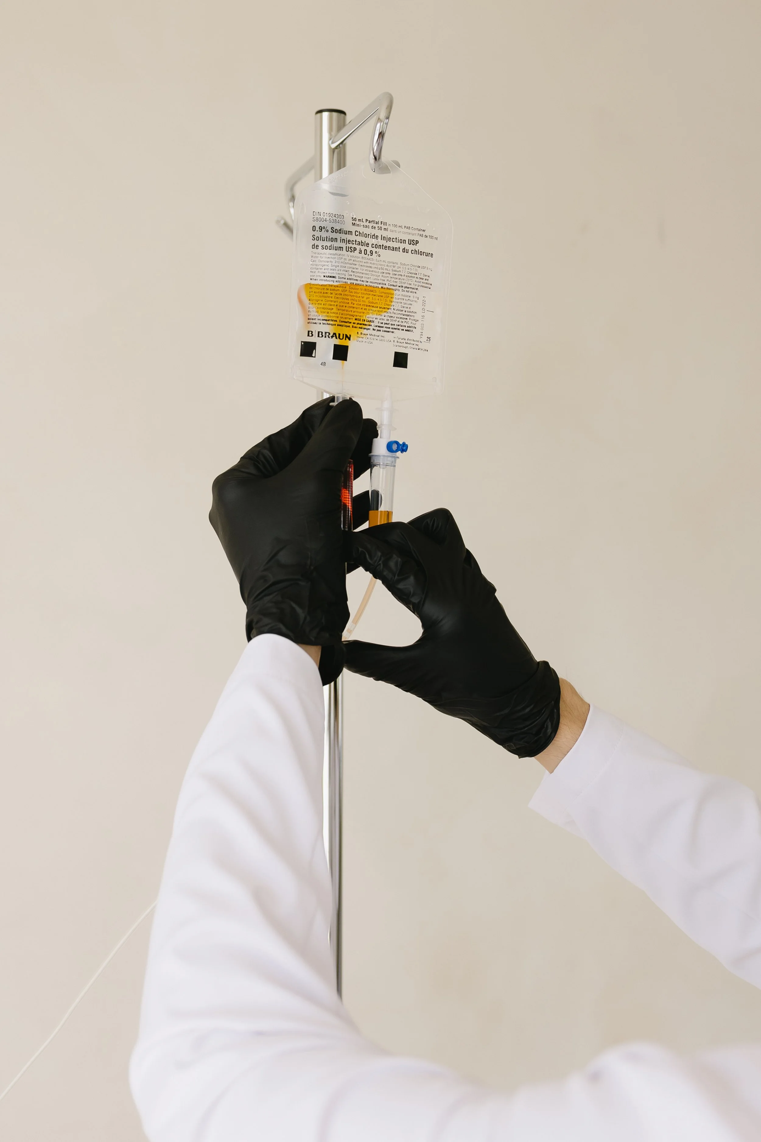 A healthcare professional wearing black gloves and white coat handling an intravenous medication bag with yellow label hanging on a metal IV pole.