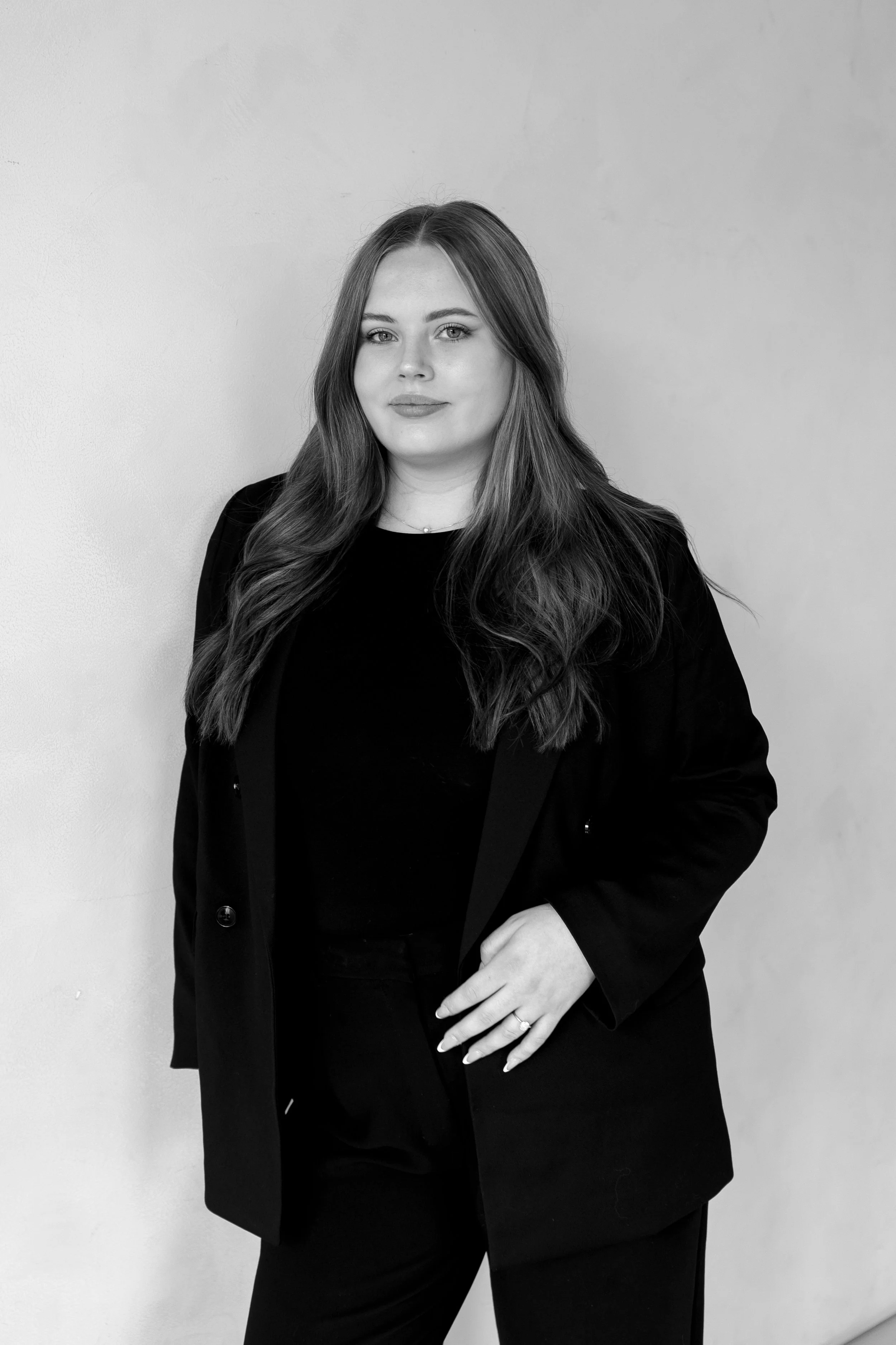 A confident woman with long, wavy hair, wearing a dark blazer over a top, standing against a plain wall.