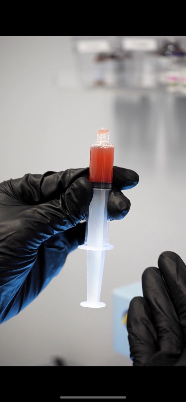 A healthcare professional wearing gloves holding two blood collection tubes, one with red blood and the other with orange fluid, over a laboratory analyzer display.