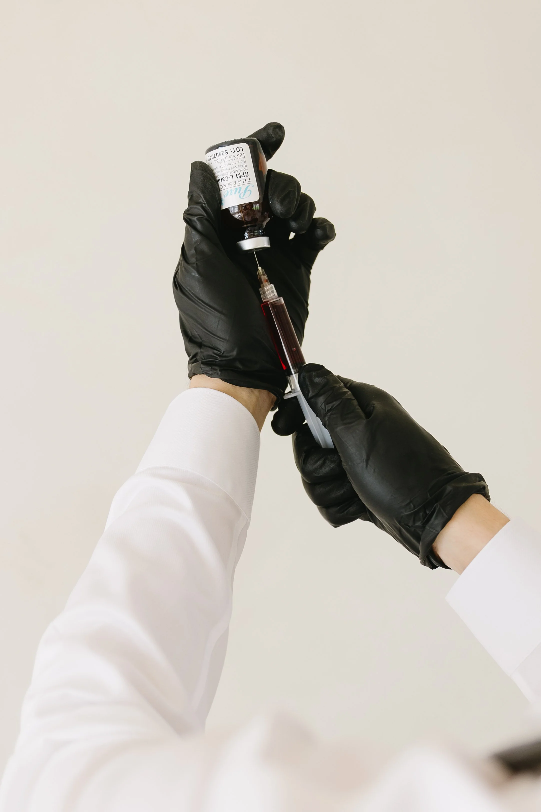 A healthcare professional wearing black gloves and a white coat prepares a syringe with a dose of medication from a small medicine vial.