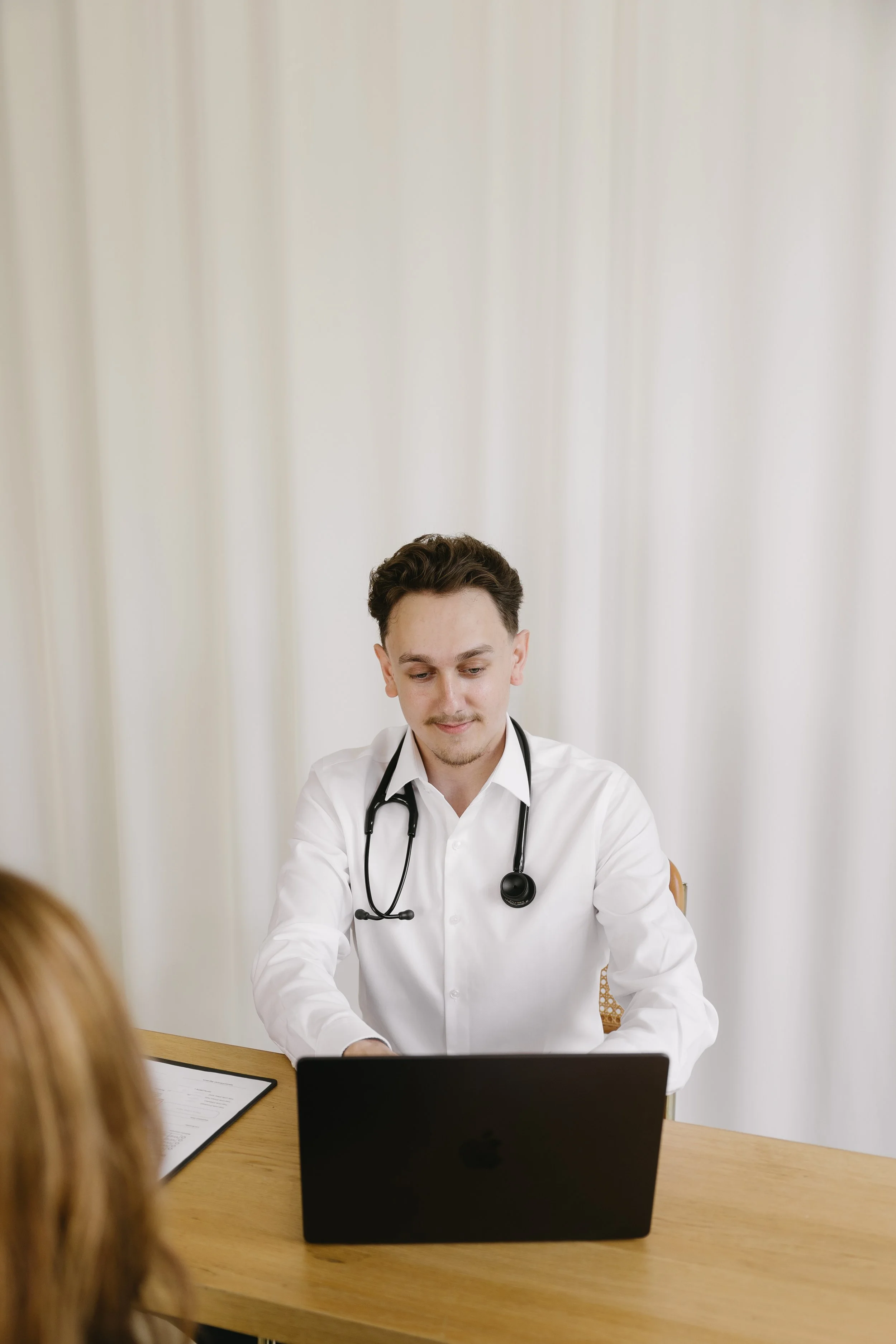 A male doctor with a stethoscope around his neck sitting at a desk with a laptop, talking to a patient.