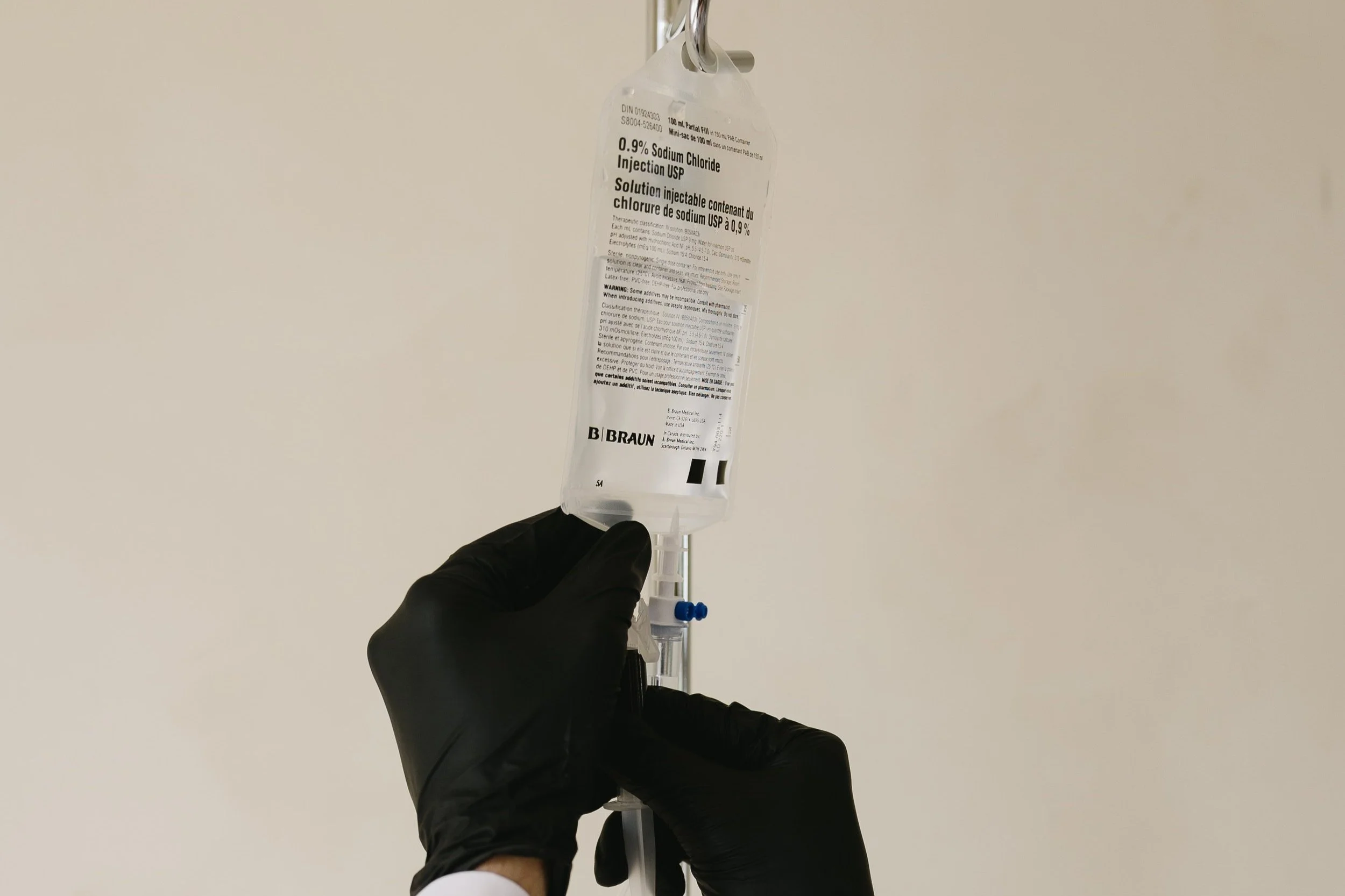 A person wearing black gloves holding a syringe connected to a bag of saline solution labeled '0.9% Sodium Chloride Injection USP' with the brand B. Braun, against a plain white background.