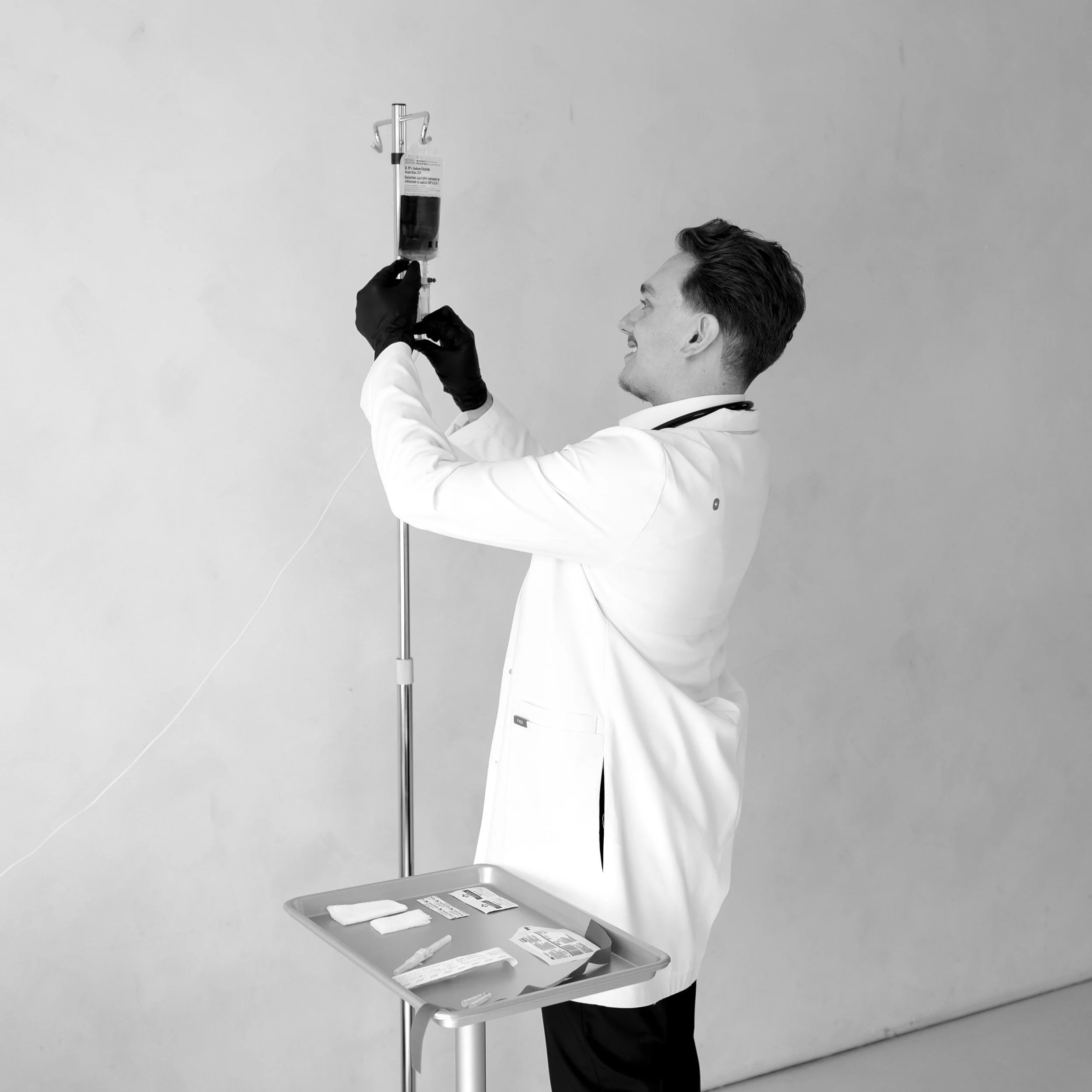 A smiling male doctor in a white coat prepares a syringe with medicine or fluid, standing next to an IV stand with a bag, and a tray of medical supplies.