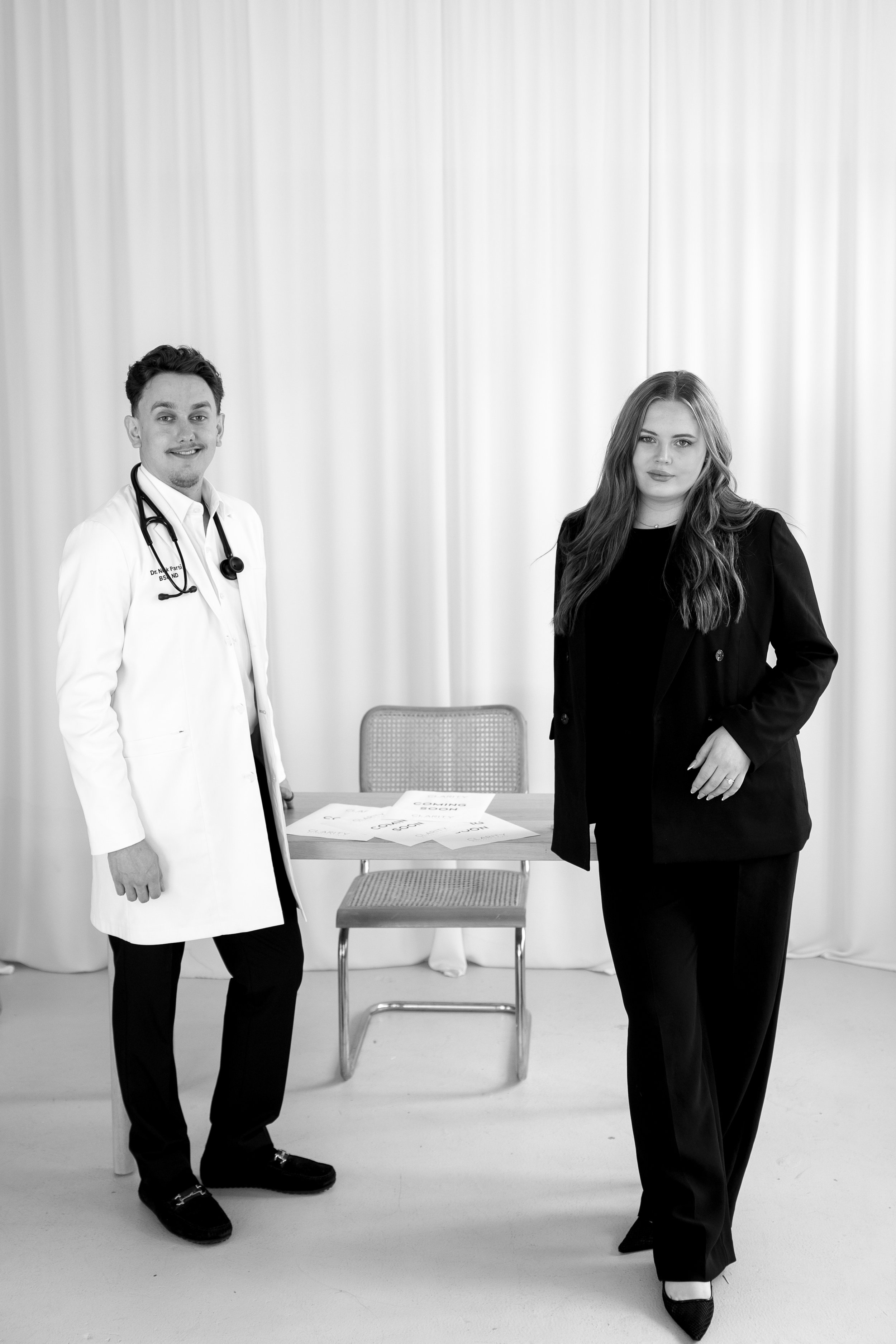 Black and white photo of a male doctor with a stethoscope around his neck and a female patient or professional standing in a room with curtains and a chair behind a table with papers.