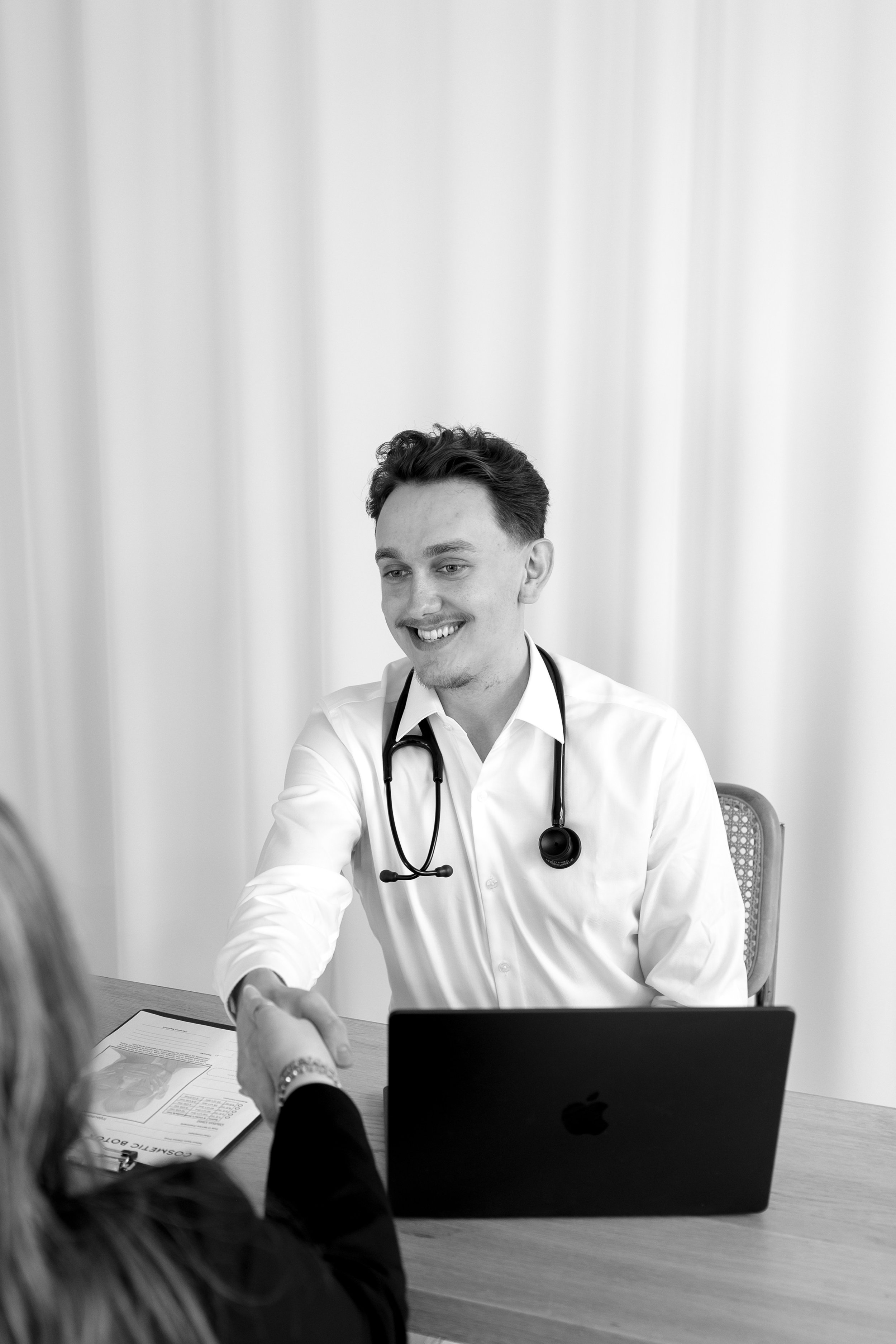A male doctor with a stethoscope around his neck smiling and shaking hands with a patient in an office setting.