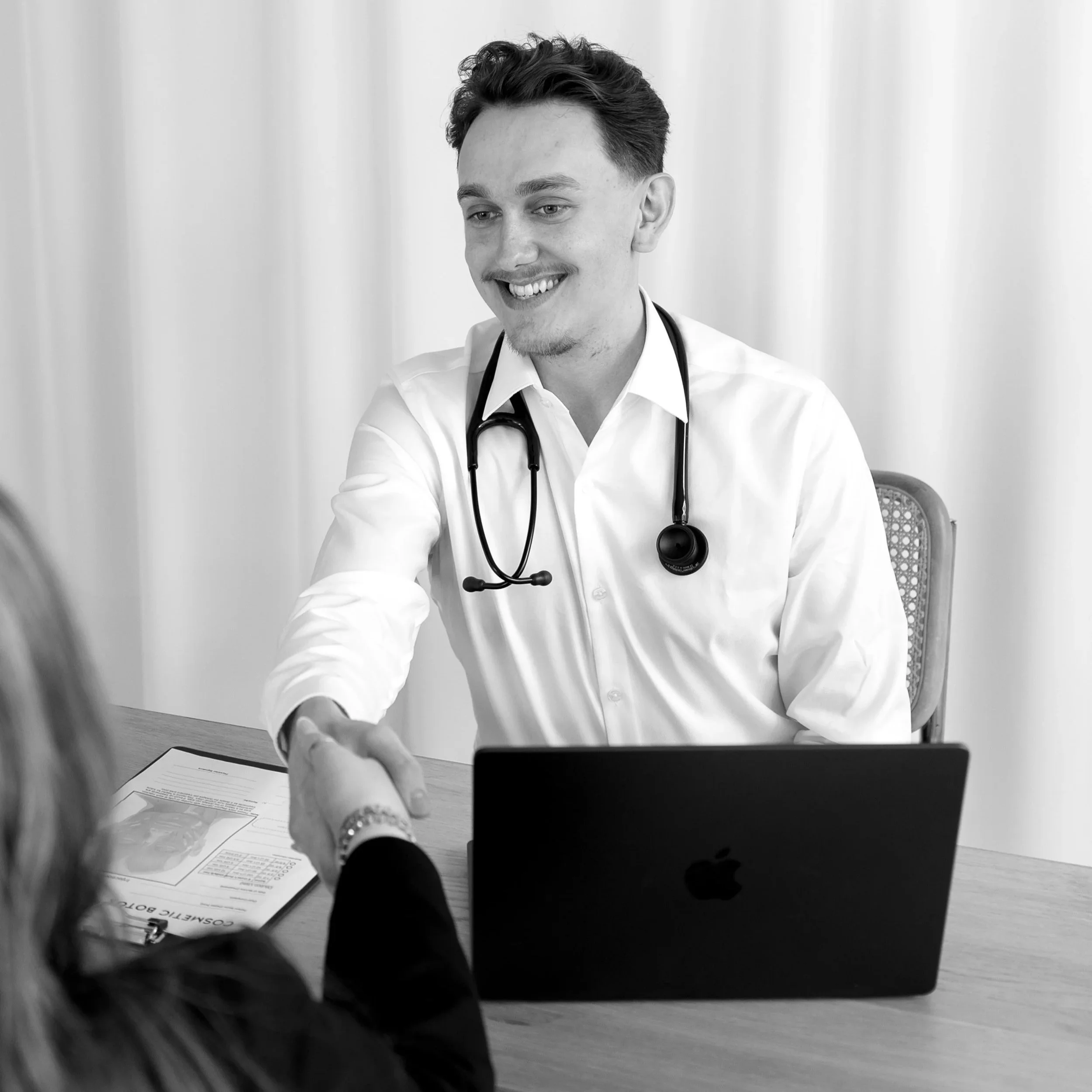 A male doctor sitting at a desk, smiling, holding a clipboard with a skin care chart, and pointing at it. There is a laptop on the desk and a woman with blonde hair in the foreground.