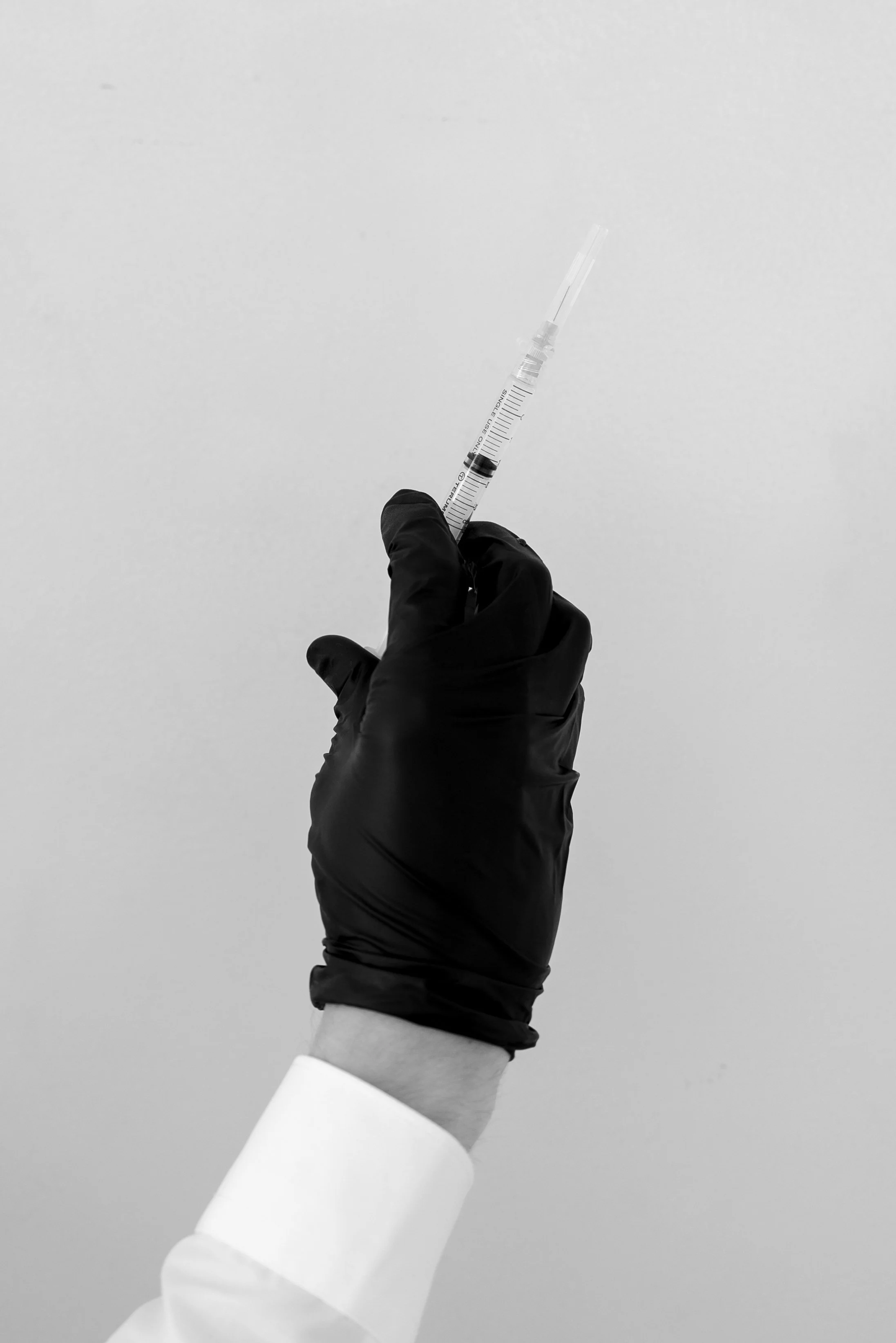 A hand wearing a black glove holding a syringe with a needle pointing upward against a plain background.