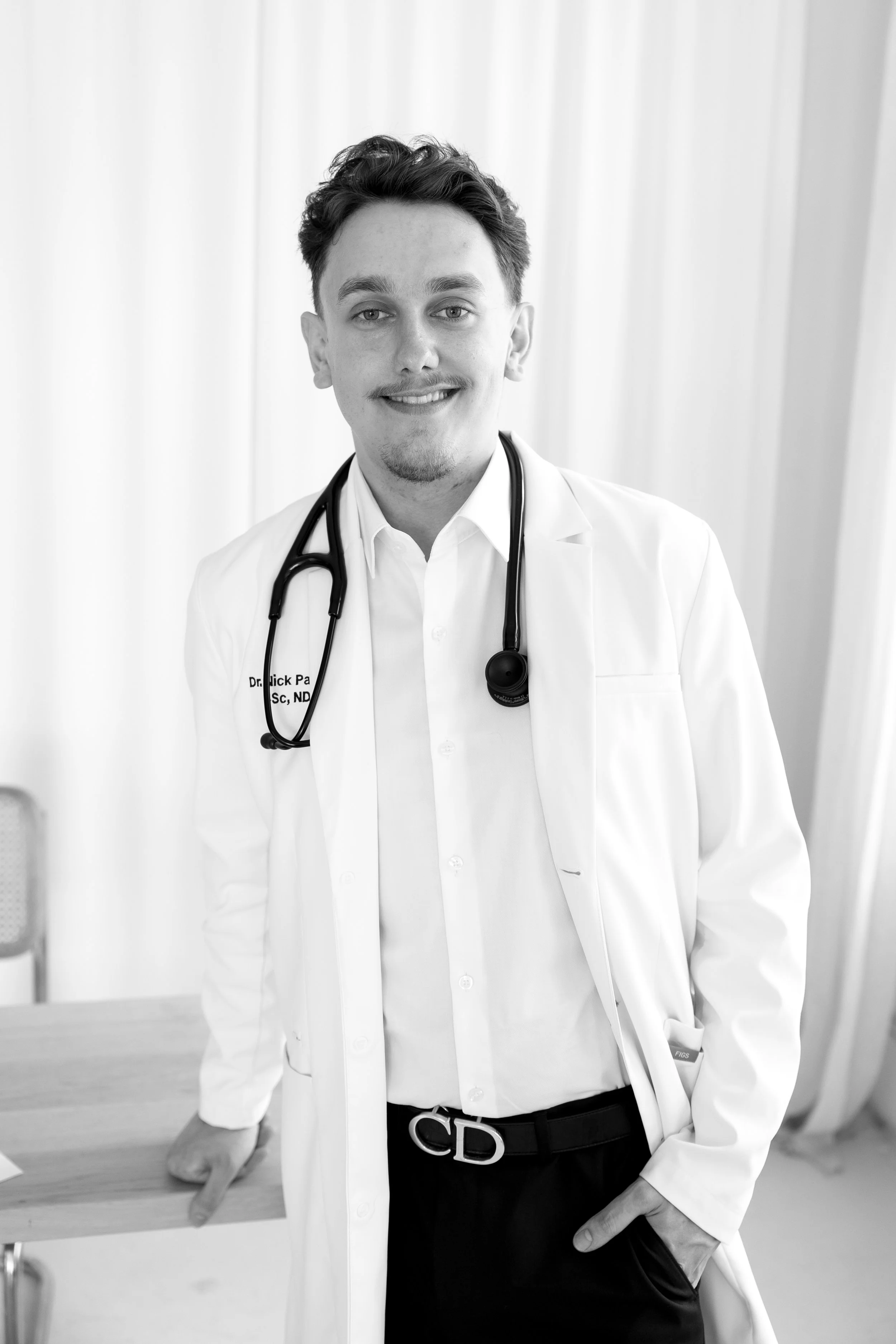 Black and white photo of a young male doctor wearing a white coat and stethoscope, smiling, standing in a clinical setting.