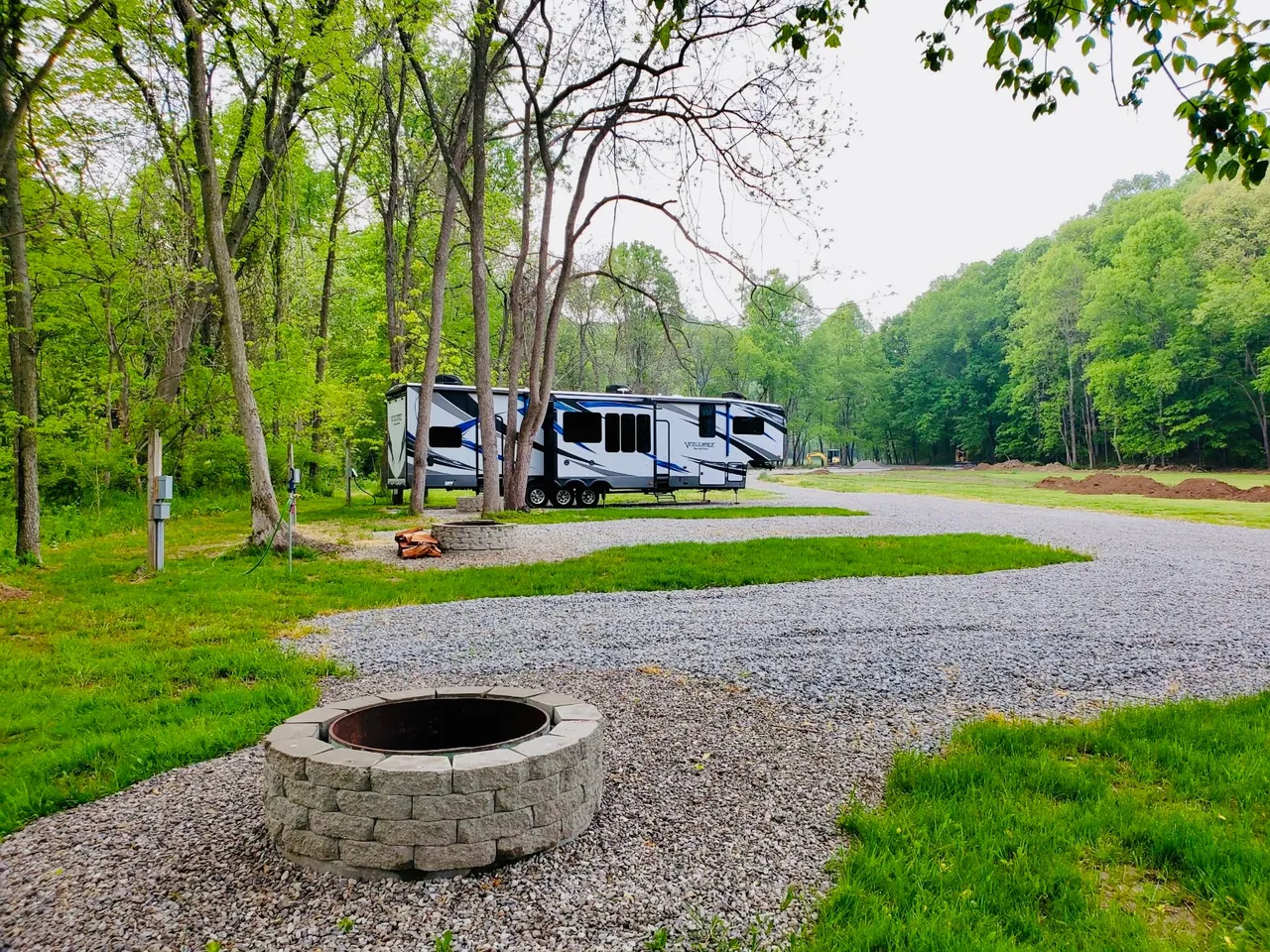 View of a camping site with a modern travel trailer parked on a gravel pad amidst green trees, a fire pit in the foreground, and a grassy area with forest in the background.