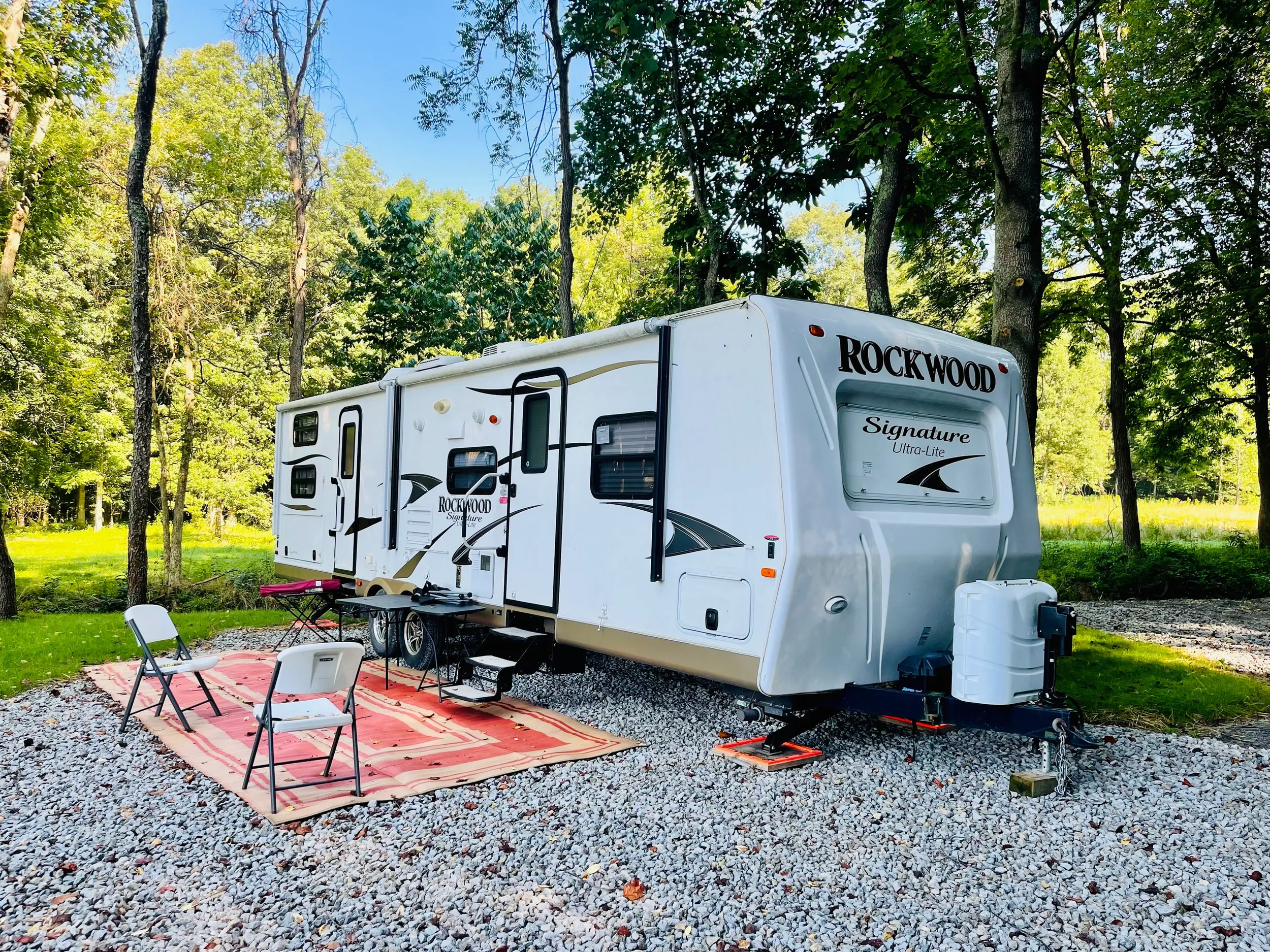 Travel trailer camper in a wooded area with outdoor seating on a red and beige rug, surrounded by green trees and grass.
