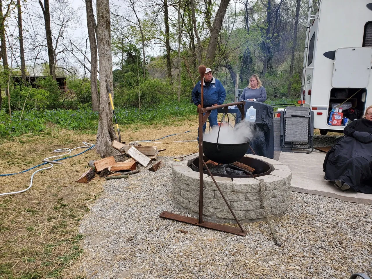 People gathering outdoors around a fire pit with a large black pot hanging above, cooking or boiling something, on a gravel surface near a tree, with a camper trailer and trees in the background.