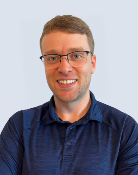 A man smiling with glasses, wearing a navy blue polo shirt, against a light background.