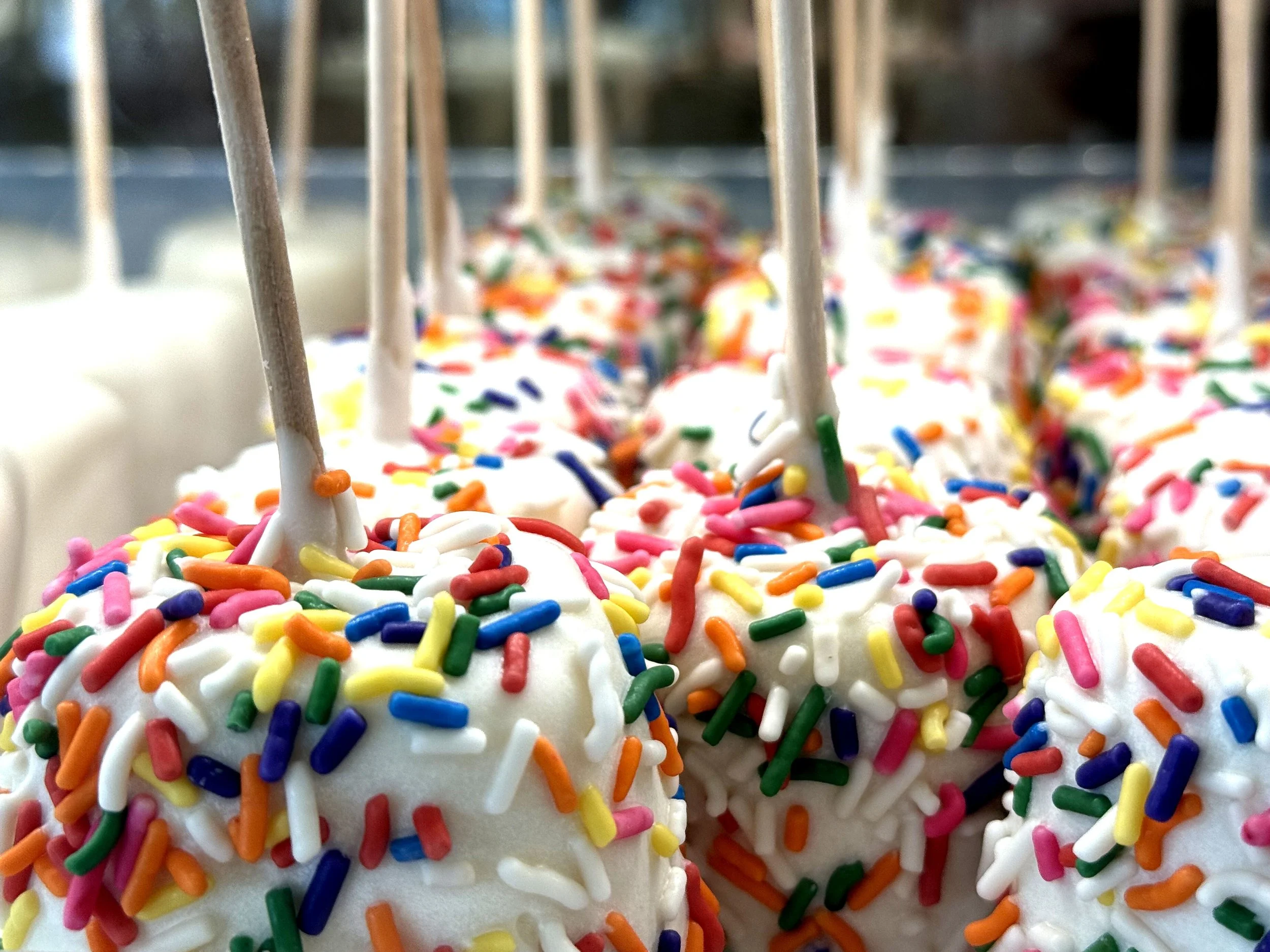 Close-up of white cake pops with colorful rainbow sprinkles and wooden sticks. Formerly Yogurtini KC.