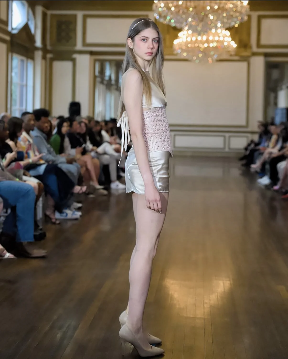 A young woman walking on a runway during a fashion show, with an audience seated on both sides, in an elegant room with chandeliers and wooden floors.