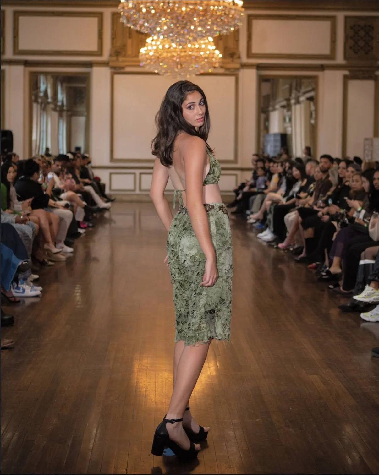 A fashion model walking down the runway at a fashion show, wearing a green lace dress and black high heels, with an audience seated on either side of the runway in an elegant room with chandeliers.