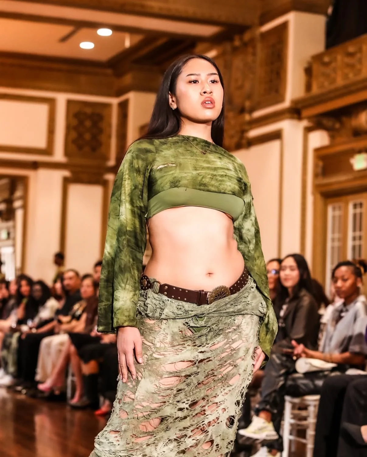 A woman walking on a fashion runway during a fashion show, wearing a distressed green crop top and a ripped skirt with a belt, with an audience watching.