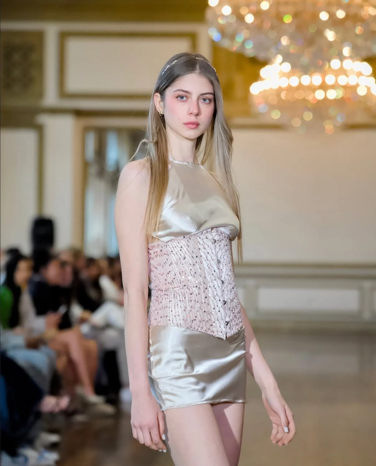 A young female model walking down a runway in a fancy, shiny outfit during a fashion show, with an audience watching in the background and a chandelier overhead.