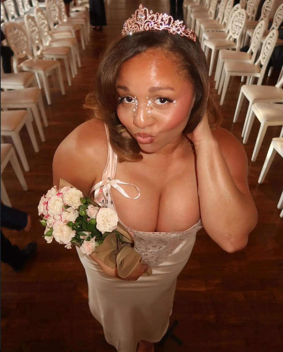 Woman wearing a tiara and dress, holding a bouquet of flowers, in a decorated room with white chairs