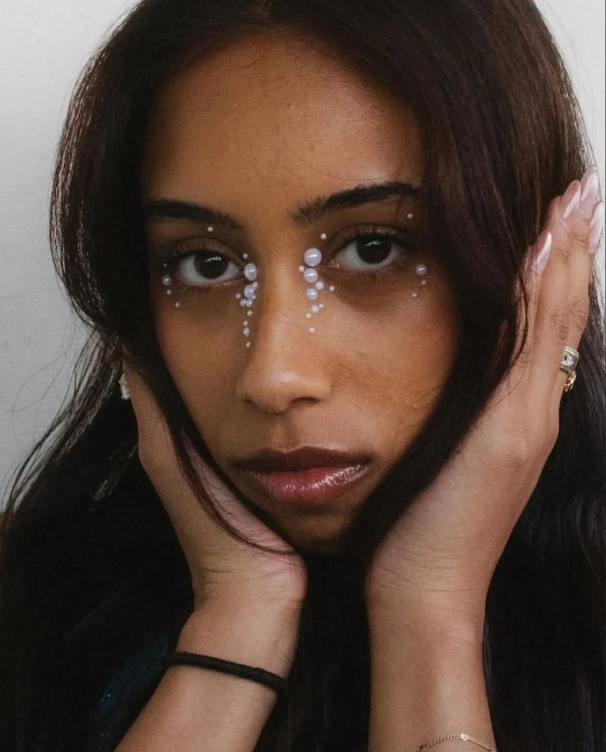A woman with dark hair and brown skin rests her face on her hands, with decorative white pearl-like beads around her eyes.