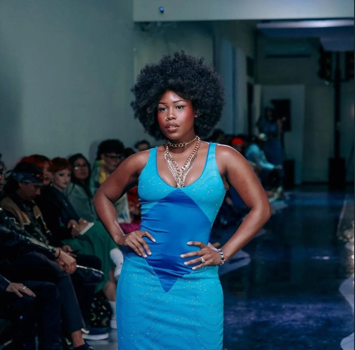 A woman walking on a runway during a fashion show, wearing a blue dress and layered gold jewelry with an audience in the background.